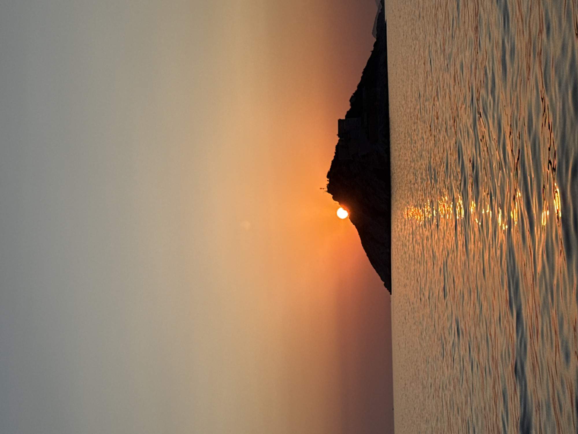 This is a vertical, eye-level, full-frame photograph taken at sunset in Mirišta, Montenegro. The image captures a serene seascape with a silhouetted island in the distance. The sun, a bright orange orb, is setting behind the island, casting a warm glow across the sky and water. The sky transitions from a soft gray at the top to a vibrant orange near the horizon. The water in the foreground reflects the sunset's colors, creating shimmering streaks of light on its surface. The island, with its rugged topography, is almost entirely in shadow, but a few structures are discernible on its peak. The overall mood is tranquil and picturesque.