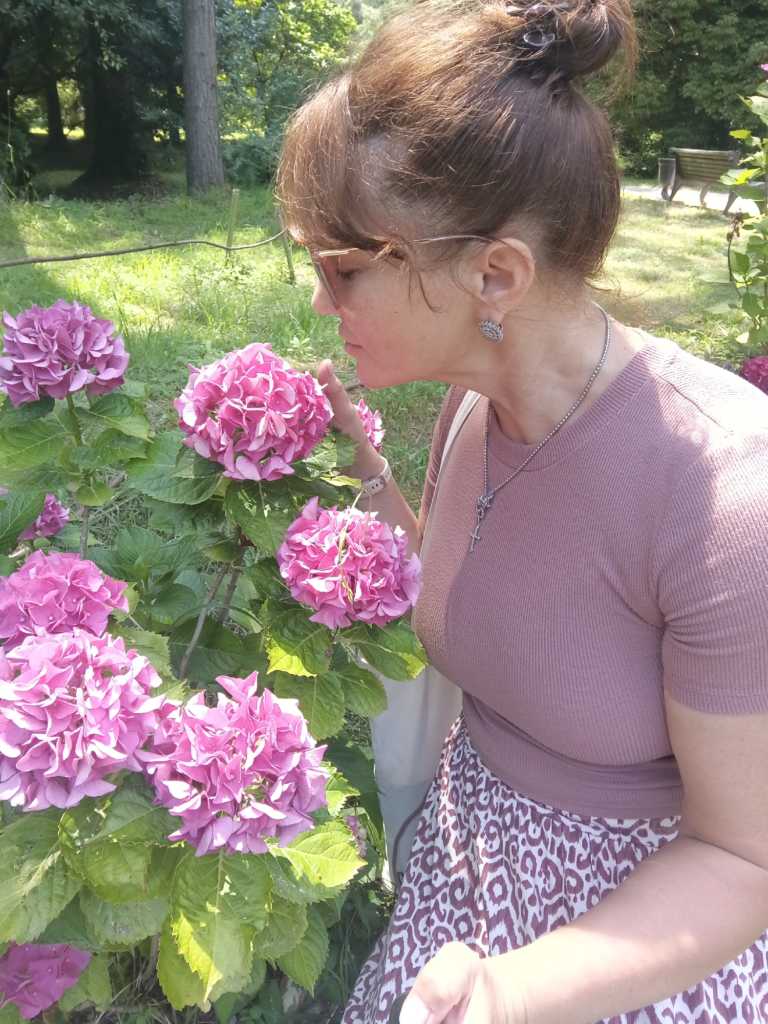 An adult female with brown hair pulled up in a bun and wearing glasses is looking at a cluster of pink hydrangeas. She is wearing a mauve-colored ribbed t-shirt and a patterned skirt. A silver necklace with a cross pendant hangs around her neck. The scene takes place outdoors in a grassy area with trees in the background, suggesting a park or garden setting. The sun is shining, casting shadows on the ground. There is a park bench visible in the background. The location is identified as Bestuzhevskoye, Russia.
