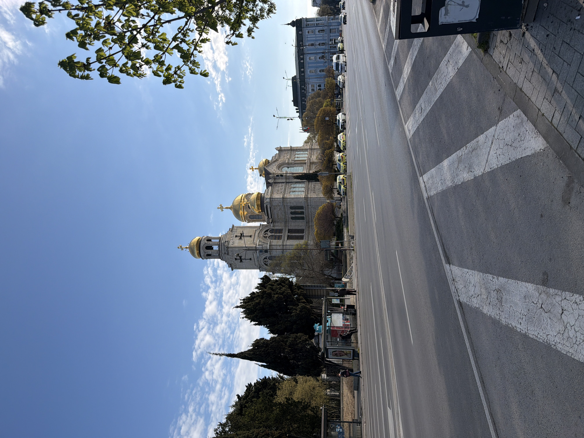 The image shows a street scene in Varna, Bulgaria, featuring the Cathedral of the Assumption of the Virgin. The cathedral, with its golden domes, stands to the left of a wide, multi-lane road. A line of trees partially obscures the base of the cathedral. A blue and white building stands in the distance beyond the cathedral. A bus stop with people waiting is located along the side of the road. A crosswalk cuts across the right side of the road, leading to a sidewalk. The sky is blue with scattered clouds, and some tree branches are visible in the upper left corner of the image. Cars are parked along the right side of the road and cars are present further away down the road.