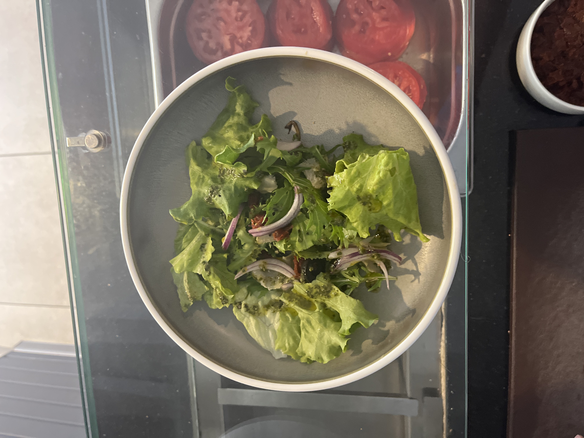 A top-down shot shows a close-up of a salad in a gray bowl. The salad is composed of large lettuce leaves, sliced red onions, and appears to be dressed with a green vinaigrette. In the background, a metal container holds sliced red tomatoes. To the right of the salad bowl, a white bowl contains a dark, chunky substance, possibly a relish or tapenade. The setting appears to be indoors, with a reflective surface below the objects, possibly glass or a polished countertop. The lighting suggests it is daytime. There is no visible text in the image.