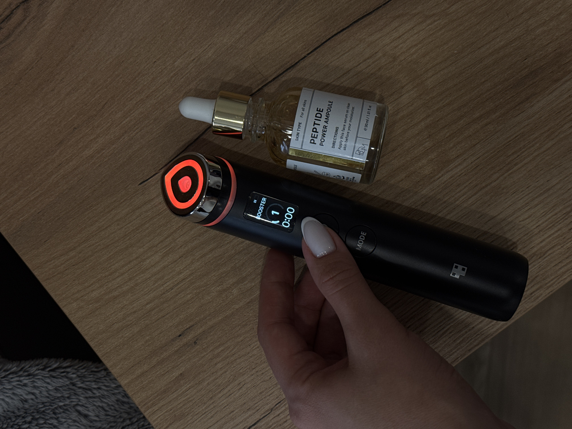 A high-angle shot depicts a cosmetic setup on a light-brown wooden surface with visible wood grain. The main elements are a black handheld skincare device, a bottle of Peptide Power Ampoule, and a female hand.
The skincare device is black with a metallic ring at the top featuring an orange light with a darker orange center. A small screen displays "BOOSTER", the number "1", and a timer set at "0:00". A "MODE" button is visible. The device is being held by a hand with a manicured white fingernail.
Next to the device sits a transparent bottle of Peptide Power Ampoule with a gold-colored dropper top. The bottle's label details information, including "SKIN TYPE: For all skins", "PEPTIDE POWER AMPOULE", directions for use, and volume "e30ml / 1.01 fl. oz".
The background consists of a wooden surface. A dark, fuzzy object is partially visible at the bottom left.