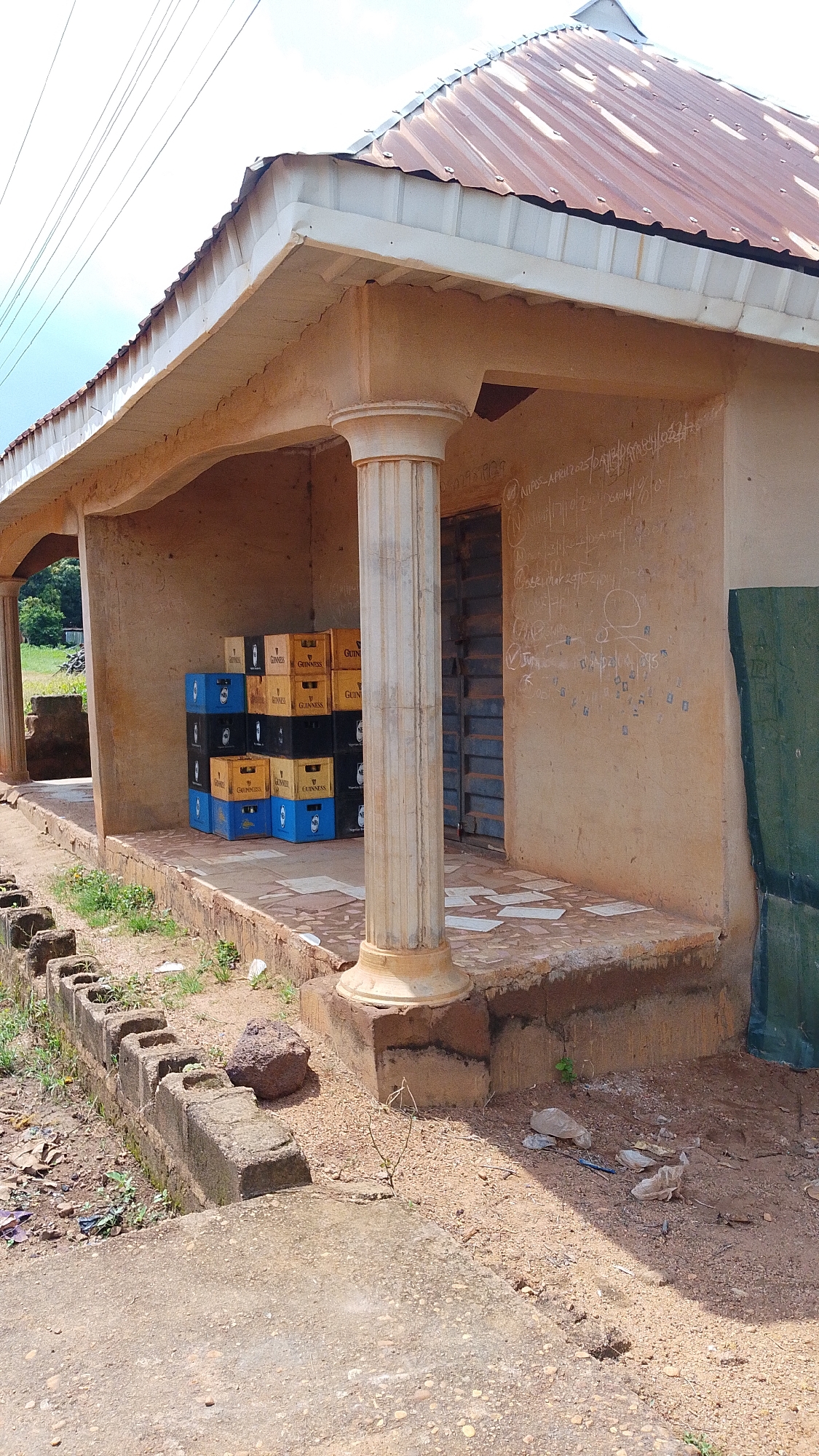 The image shows an outdoor scene in Donga, Nigeria, likely during the day with good weather. The primary subject is a small, rustic building with a corrugated metal roof and light-colored, stuccoed walls. The building has a covered porch supported by a decorative column.
On the porch, a stack of beverage crates is visible. These crates are primarily blue and yellow, and some of them bear the Guinness logo. The crates suggest a commercial establishment, possibly a small shop or a place selling drinks.
The walls of the building are marked with chalk or a similar substance, forming what appears to be handwritten notes, dates, and possibly drawings. Some legible text includes "NURDS - APEN 2015" and "30/10/19/2". The presence of these markings could indicate a place frequented by many people, or perhaps a communal bulletin board.
In front of the building, there is a concrete walkway and a raised concrete curb or planter. The ground is covered with dirt and gravel, with patches of dry grass and weeds growing in places. A few rocks are scattered on the ground, one of which is quite large. The overall setting appears to be somewhat rural or a less developed urban area, given the unpaved ground and simple construction. The scene is quiet, with no people visible, and the lighting suggests it's daytime.