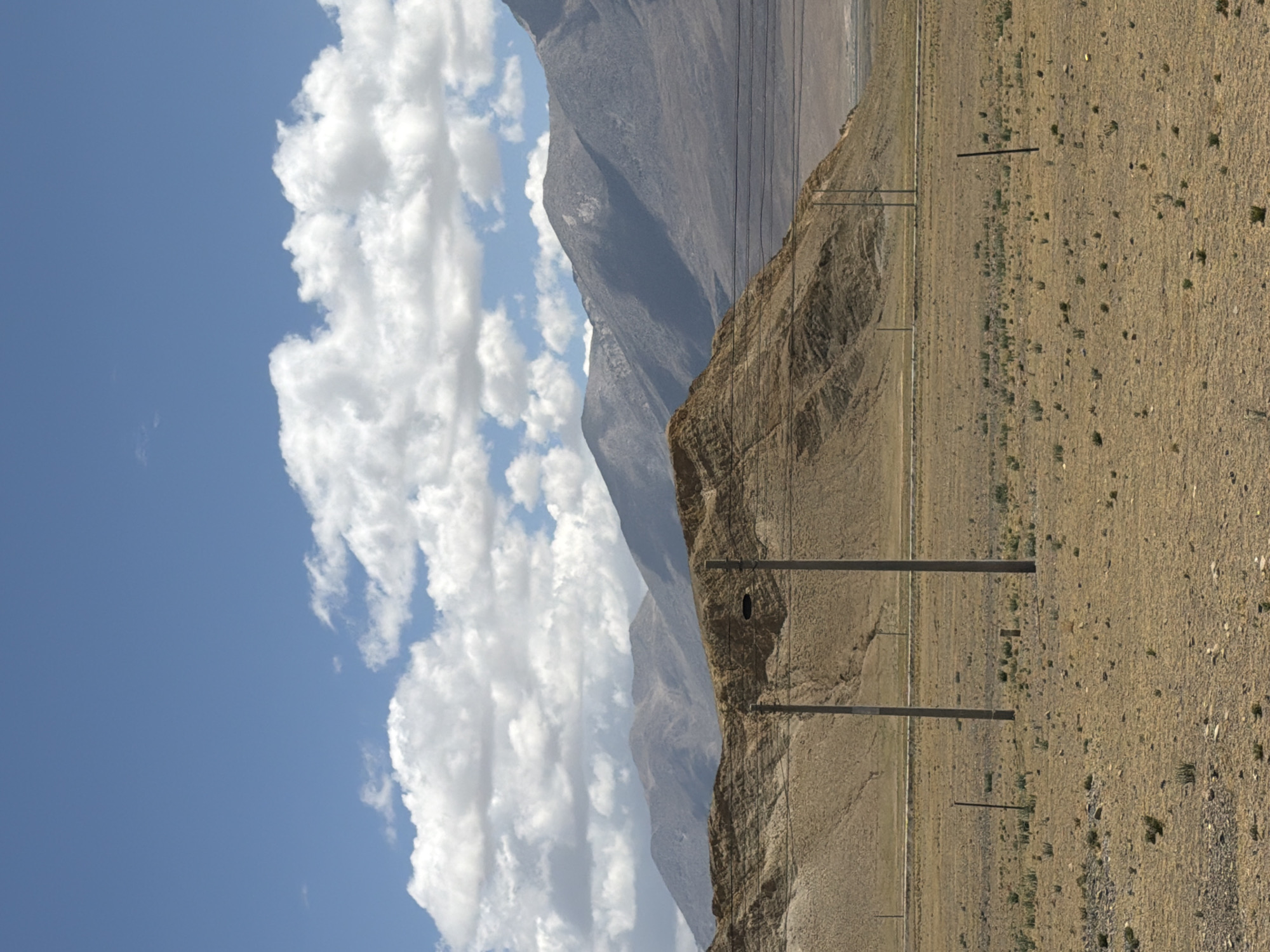 A vast, arid mountainous landscape is depicted under a bright blue sky. The upper portion of the image is dominated by the clear blue sky, interspersed with scattered, fluffy white cumulus clouds.
In the middle ground, a large, dark, rugged mountain range extends across the frame. The mountains appear barren with visible rock formations and steep slopes.
The foreground, particularly on the right and lower sections, features an expanse of dry, light brown earth, possibly a plain or roadside, strewn with small rocks and very sparse, dry vegetation.
Several tall, slender utility poles are visible, standing vertically in both the foreground and mid-ground. Wires are strung between some of these poles, traversing the scene across the mountains and sky. One specific utility pole in the mid-ground has a distinct circular dark opening or cavity near its top.
The scene suggests bright daylight and clear weather. No human or animal activity is visible. There is no visible text. The location context provided is Mainpu township, China. The image conveys a sense of remoteness and rugged natural beauty.