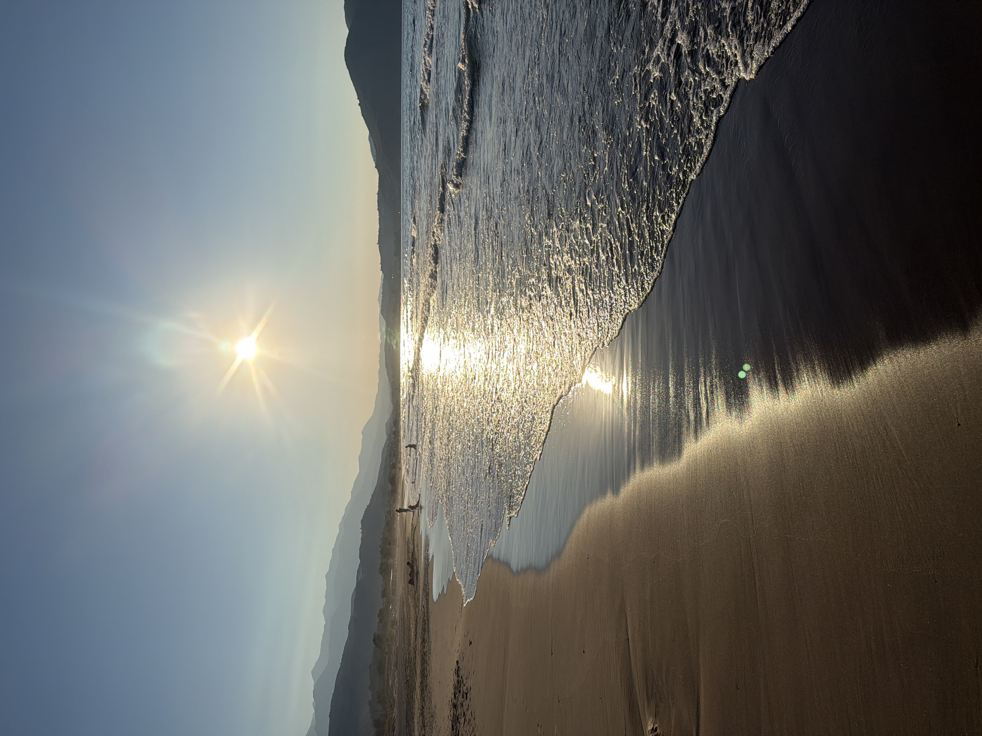 The image depicts a serene beach scene at sunset in Georgioupoli, Greece. The sun, a bright starburst, hangs low in a clear blue sky, casting a warm, golden glow across the landscape. Gentle waves lap the sandy shore, their crests shimmering with sunlight. In the distance, hazy mountains form a picturesque backdrop. A few individuals are visible on the beach and in the shallow water, their figures silhouetted against the bright horizon. The sand, ranging in color from light tan to a darker, wet sheen near the water's edge, is smooth and undisturbed, save for the receding tide. The overall impression is one of tranquility and natural beauty, characteristic of a Greek coastal town at dusk.