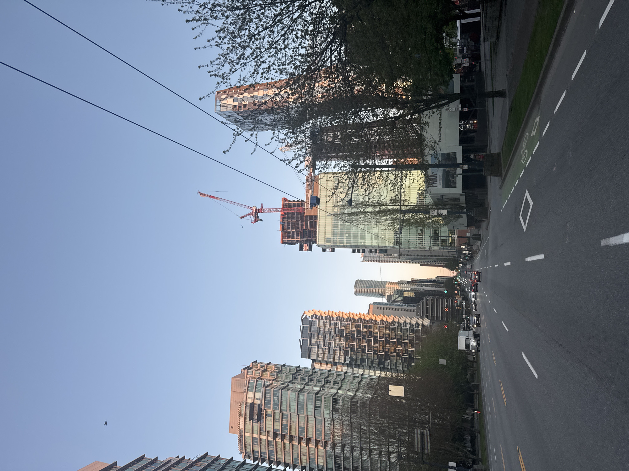 The image presents an urban street scene in Vancouver, Canada, bathed in the warm, golden light of what appears to be late afternoon or early evening. The sky is clear and blue.
A wide asphalt road runs through the center of the frame, featuring multiple lanes marked by dashed white lines and yellow double lines further down. A distinct green-painted bike lane, indicated by a white bicycle symbol, is visible on the left side of the road, bordered by a solid white line. Vehicles, including cars and trucks, are present on the road, particularly in the distance, suggesting typical city traffic.
The street is flanked by numerous modern high-rise buildings, showcasing diverse architectural styles. Many buildings feature expansive glass facades that reflect the surrounding light and structures. One building under active construction is prominent, with its exposed framework and a towering red construction crane. Another notable structure exhibits a unique checkered or patterned facade near its upper levels, and several buildings display intricate, staggered balcony arrangements. Deciduous trees, some with sparse leaves, line parts of the street.
Visible text includes "PARK" on what appears to be development banners or signs, and a "50" speed limit sign. Power lines are visible stretching across the clear sky. No individuals are clearly discernible. The scene captures the dynamic and developing nature of a modern city.