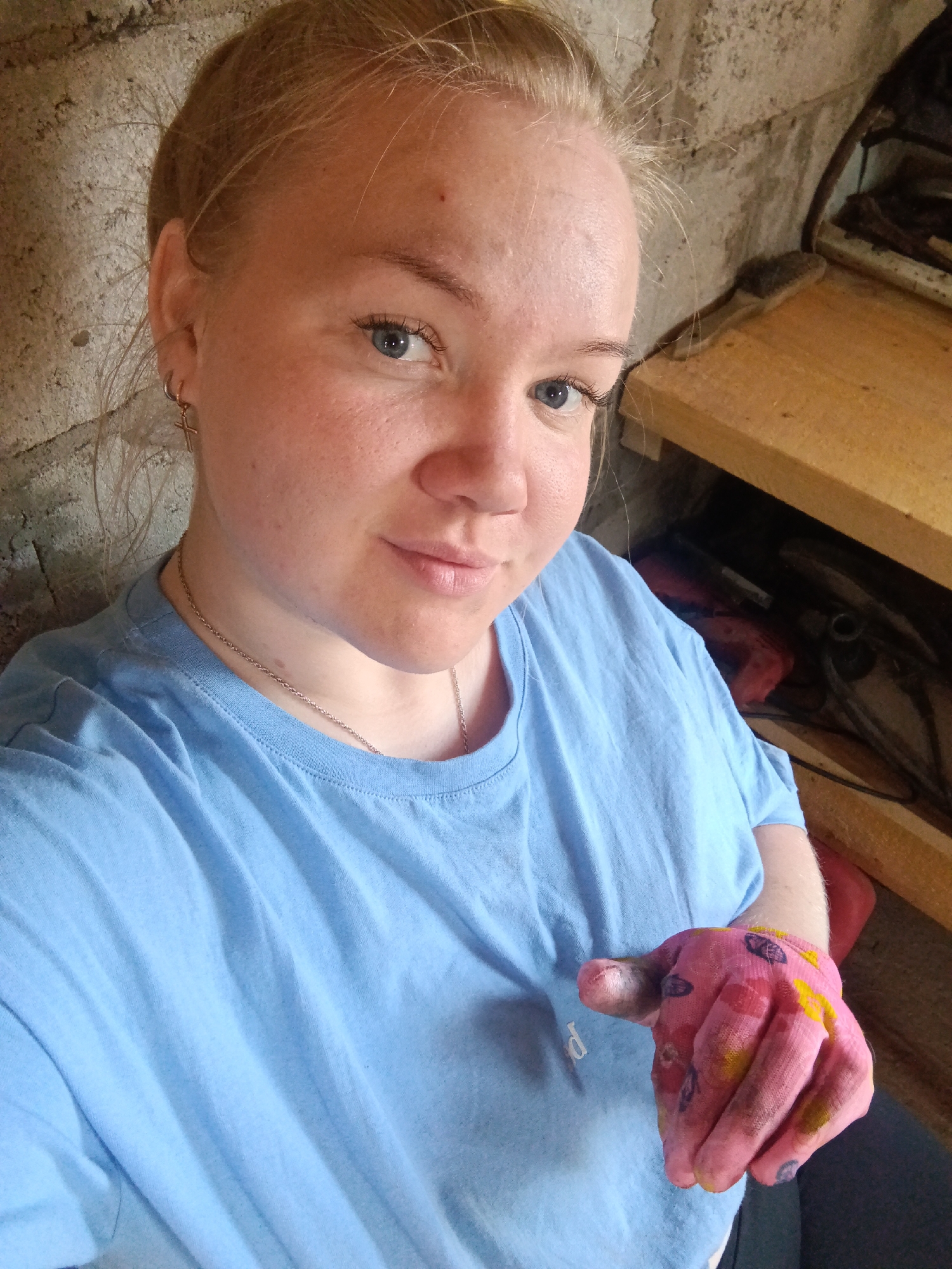 A woman with blonde hair pulled back is taking a selfie. She has blue eyes and is wearing a light blue t-shirt and a silver necklace. Her right hand is in the foreground, covered in pink, yellow, red, and black paint. She is wearing an earring that appears to be a cross on her left ear. The background shows a rough concrete wall on the left and a wooden shelf unit with miscellaneous items on the right. This scene is indoors. Фоки, Russia is not discernible from the image.