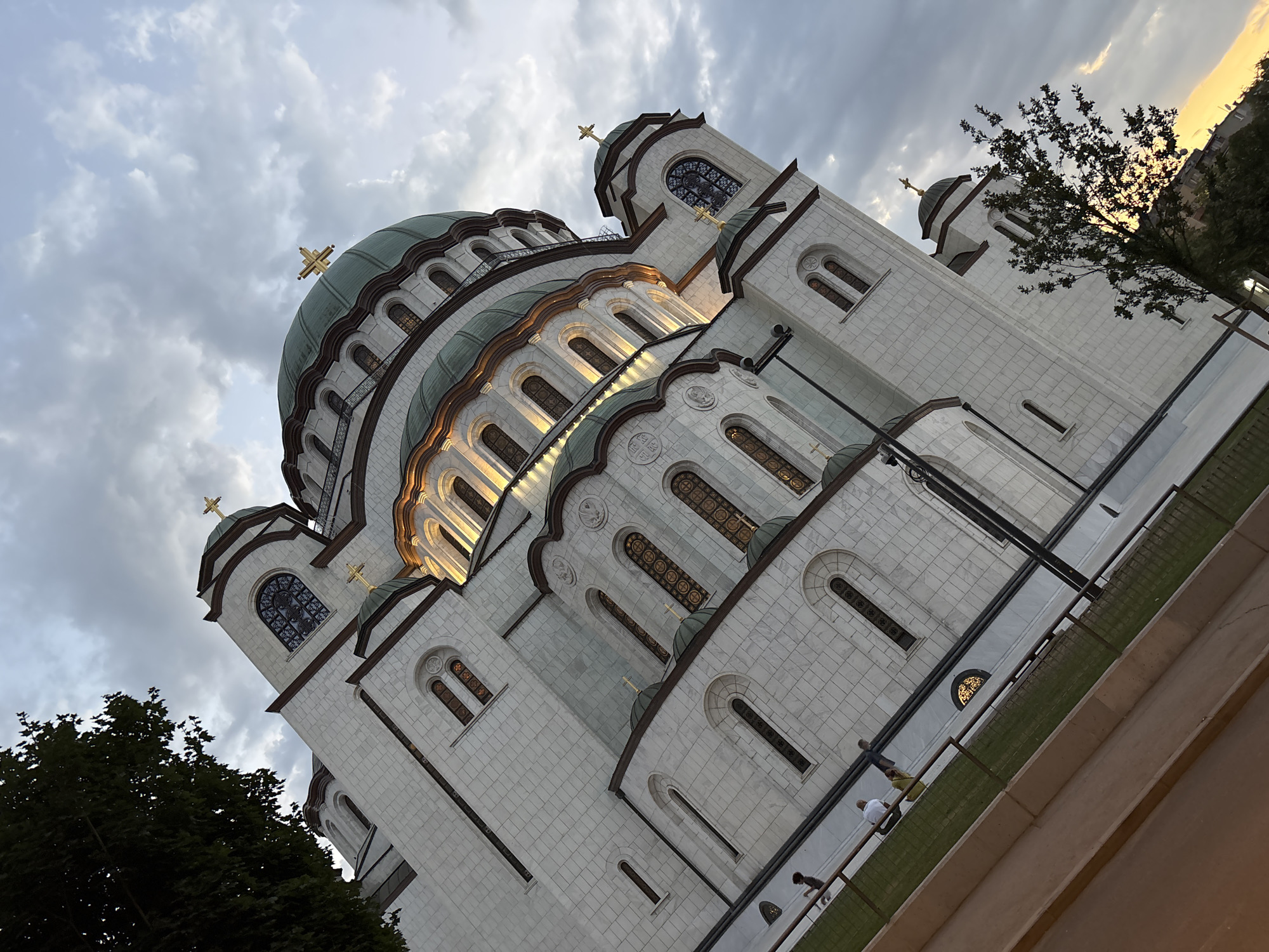 The image shows the exterior of the Church of Saint Sava in Belgrade, Serbia, under a cloudy sky. The church is a large, white stone building with multiple domes, many arched windows with decorative stained glass, and golden crosses on top of the domes. The building is illuminated from within, creating a warm glow around the windows.
In the foreground, there is a grassy area enclosed by a railing. A few people are visible walking along a paved path next to the grassy area. To the left of the church, a large tree with dark green leaves is visible. To the right and in the background, the setting sun casts a warm yellow and orange glow, partially obscured by buildings and trees. The camera angle is tilted, giving a dramatic perspective of the church.