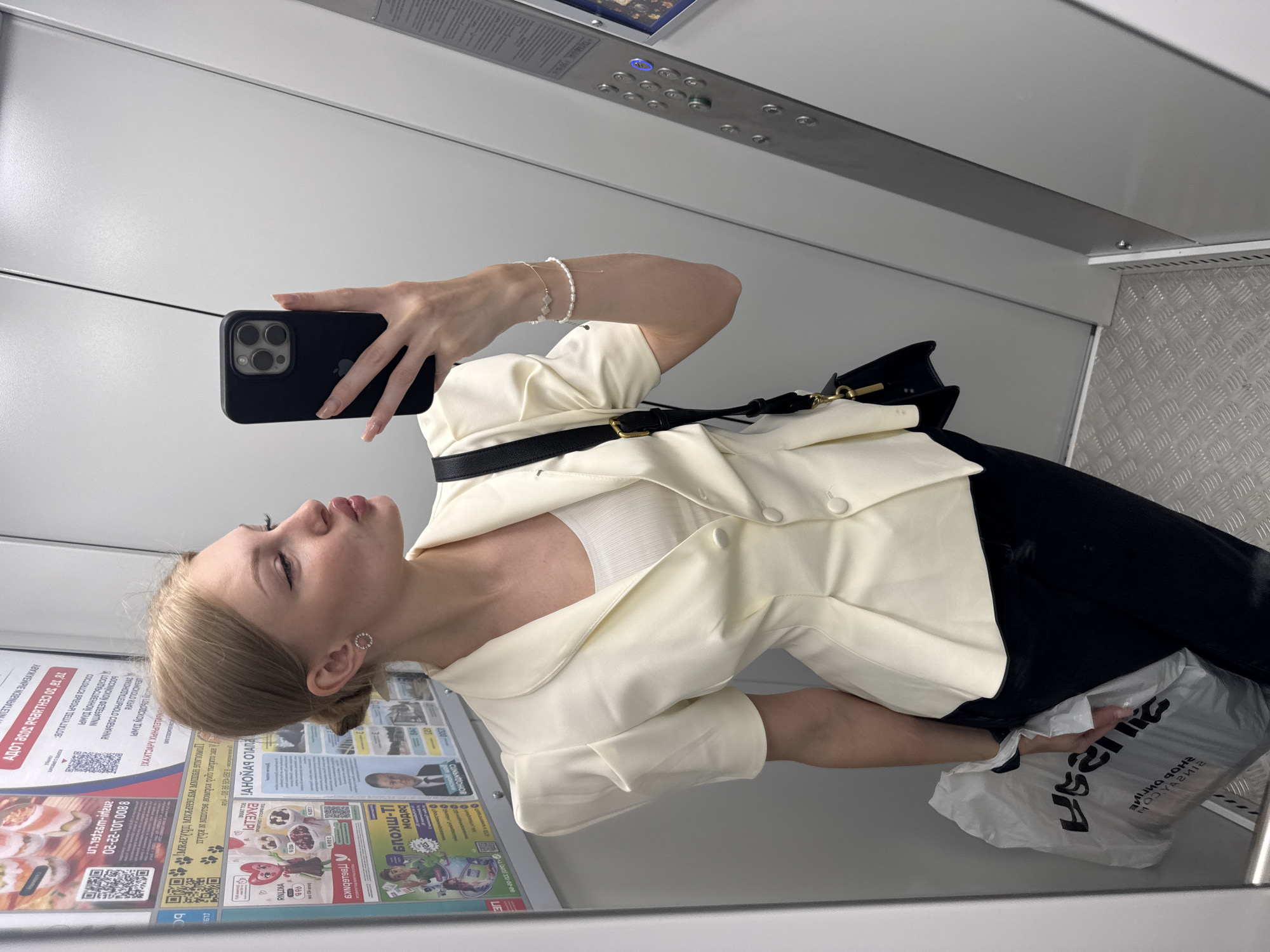 In an elevator in Perm, Russia, a young Caucasian woman with blonde hair styled in a bun takes a mirror selfie. She is wearing a light yellow, short-sleeved blazer with padded shoulders over a white tank top and black pants. A black crossbody bag with gold hardware hangs from her shoulder. She is holding a smartphone in her right hand, angled to capture the reflection. In her left hand, she holds a white plastic shopping bag. The elevator walls are gray, and the floor is made of metal diamond plate. Posters are visible on the wall behind her.