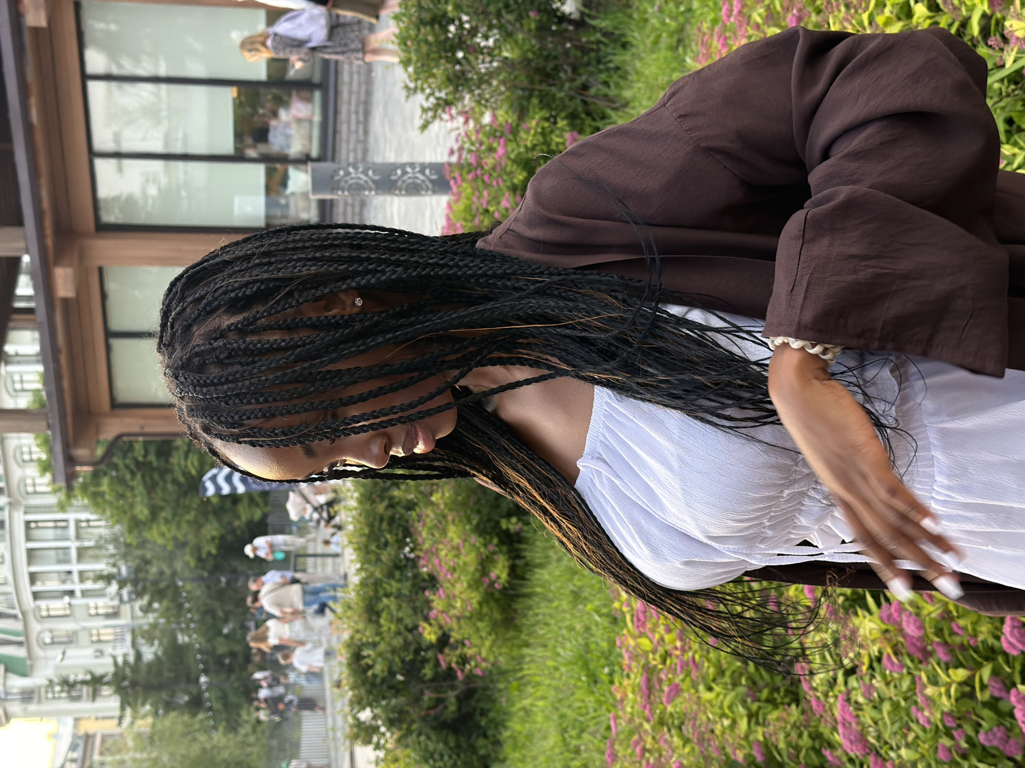 The image shows a young Black woman with long, braided hair, wearing a white off-the-shoulder top and a brown open-front jacket. She is standing in front of a backdrop of lush green grass and pink flowers, with a building and other people visible in the blurred background. The lighting suggests it is daytime, possibly late afternoon due to the warm tones.
While the image depicts an outdoor scene in a city environment, there is no specific visual information within the image itself to confirm the location as Kazan, Russia. The architectural style of the building in the background is not distinctly identifiable as Russian. The people in the background are too blurred to discern specific cultural attire or features that would pinpoint the location. Therefore, without external information, it is impossible to confirm this location.