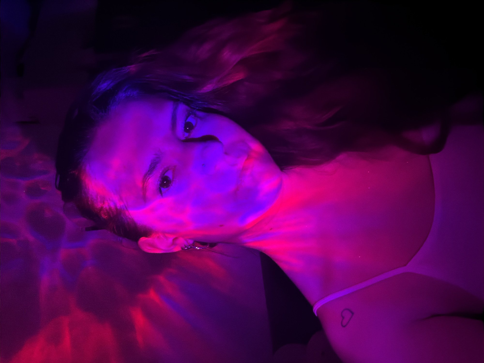 A person is lying down, illuminated with a blend of pink and purple light. The light creates a shimmering effect on the surface near the person's head. The person has long hair, visible facial features, and is wearing a garment with a thin strap. A small heart tattoo is visible on the person's upper back. The environment appears dark beyond the illuminated areas. There is no indication of the location being Vozdovac Urban Municipality, Serbia.