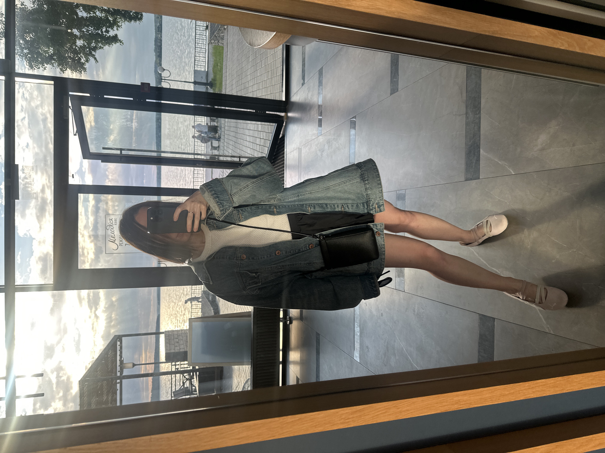 A woman is taking a mirror selfie in a commercial establishment, possibly a cafe or restaurant, located in Votkinsk, Russia, with a view of the Votkinsk Reservoir. The woman is wearing a denim jacket over a cream-colored top and dark shorts, with light pink flat shoes. She is holding a black smartphone in her right hand and a small black crossbody bag is slung over her shoulder. Reflected in the mirror behind her, the exterior shows a body of water, a covered outdoor seating area, and several people walking or sitting by the waterfront. The interior floor is tiled with a grey marble pattern. The overall lighting suggests it is late afternoon or early evening, with the sun casting rays through the clouds.