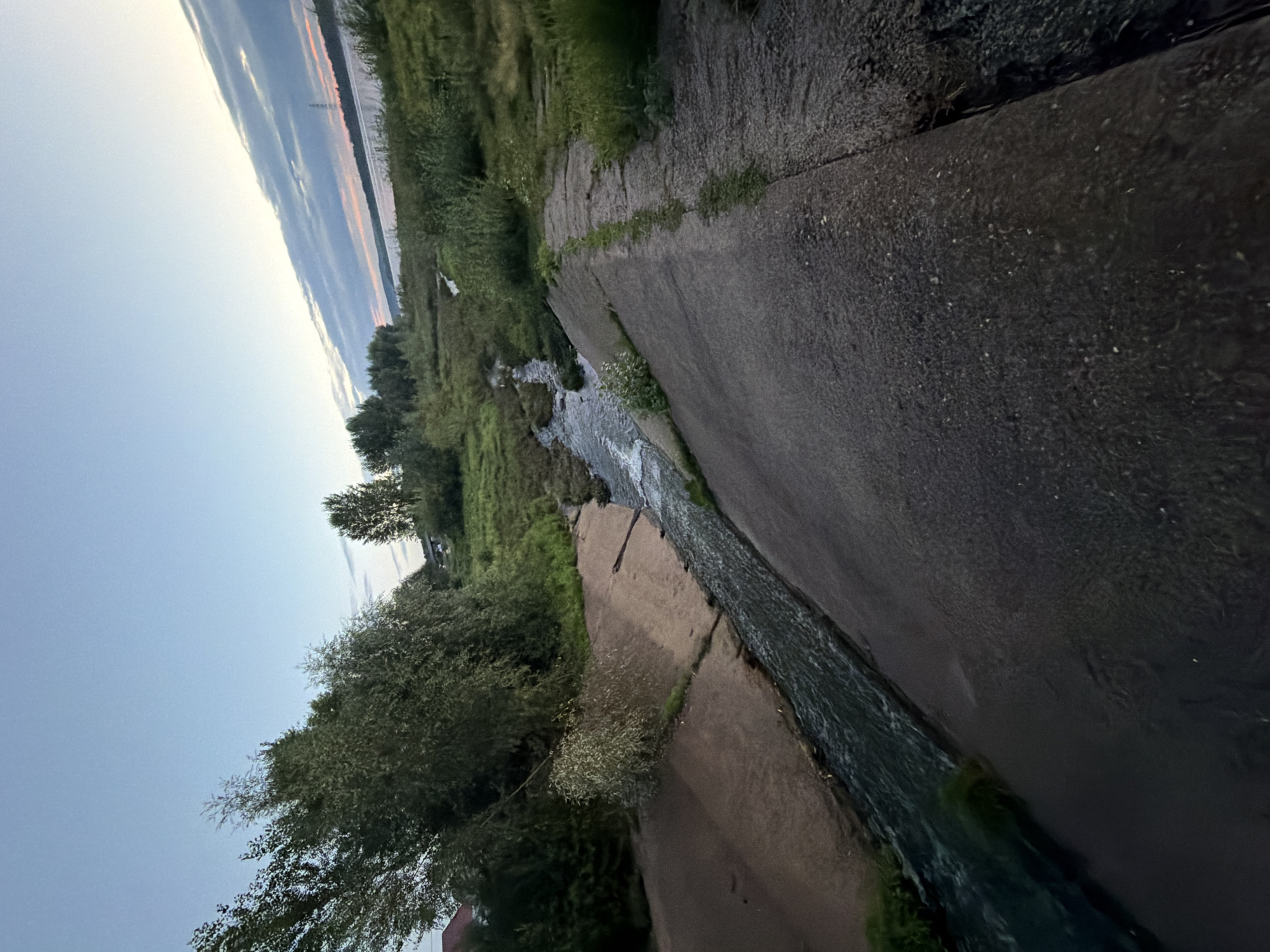 An outdoor, medium shot, low-angle photograph shows a drainage ditch or canal with water flowing through it. The canal has concrete walls on one side and earthen banks on the other. Lush green vegetation, including trees and bushes, grows on the banks and surrounding landscape. In the background, a wide body of water, likely the Kama River, is visible under a twilight sky with wispy clouds. The scene appears to be a natural or semi-natural area within the city of Chaikovsky, Russia, possibly a man-made waterway designed to manage water flow. There are no people or animals present in the image. The lighting suggests it is either dawn or dusk.