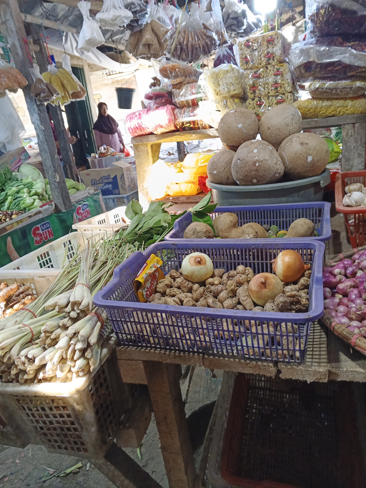 This is a full-shot, eye-level, outdoor photograph of a market stall in Warungkiara, Indonesia. The stall is filled with various produce, including coconuts, onions, ginger, lemongrass, and packaged snacks. A woman wearing a headscarf is visible in the background, standing near the entrance of the stall. The stall appears to be made of wood and has a rustic, open-air design. Sunlight streams in from the top, illuminating the scene and casting shadows. The ground is dirt and debris, indicative of a bustling market environment.