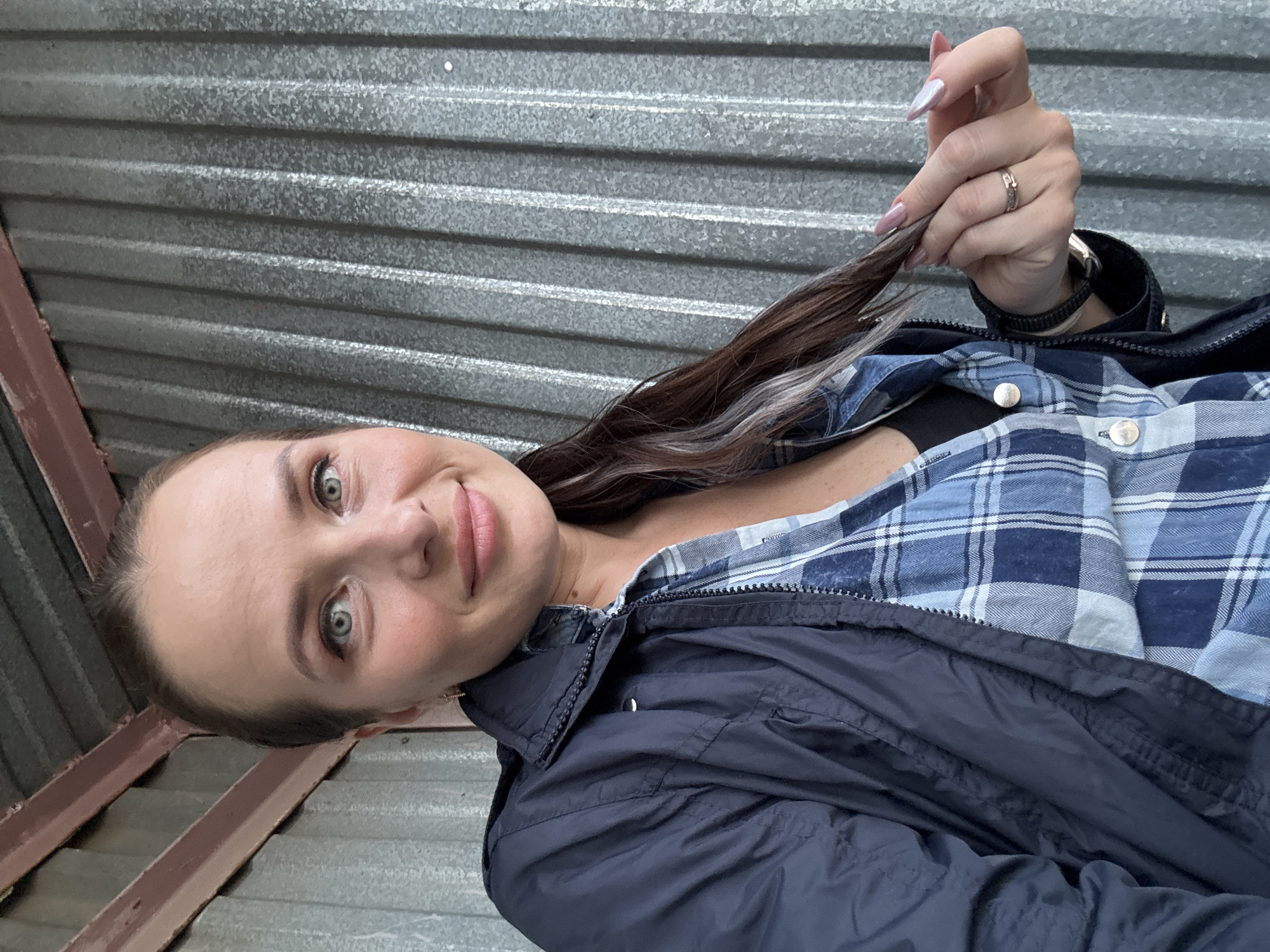 A woman is pictured in a close-up shot, holding a strand of her hair in her right hand. She is wearing a blue plaid shirt and a dark navy jacket. A ring is visible on her right ring finger, and she is wearing a watch on her left wrist. The background appears to be a corrugated metal wall. There are no indicators to specify the location in Chaikovsky, Russia.