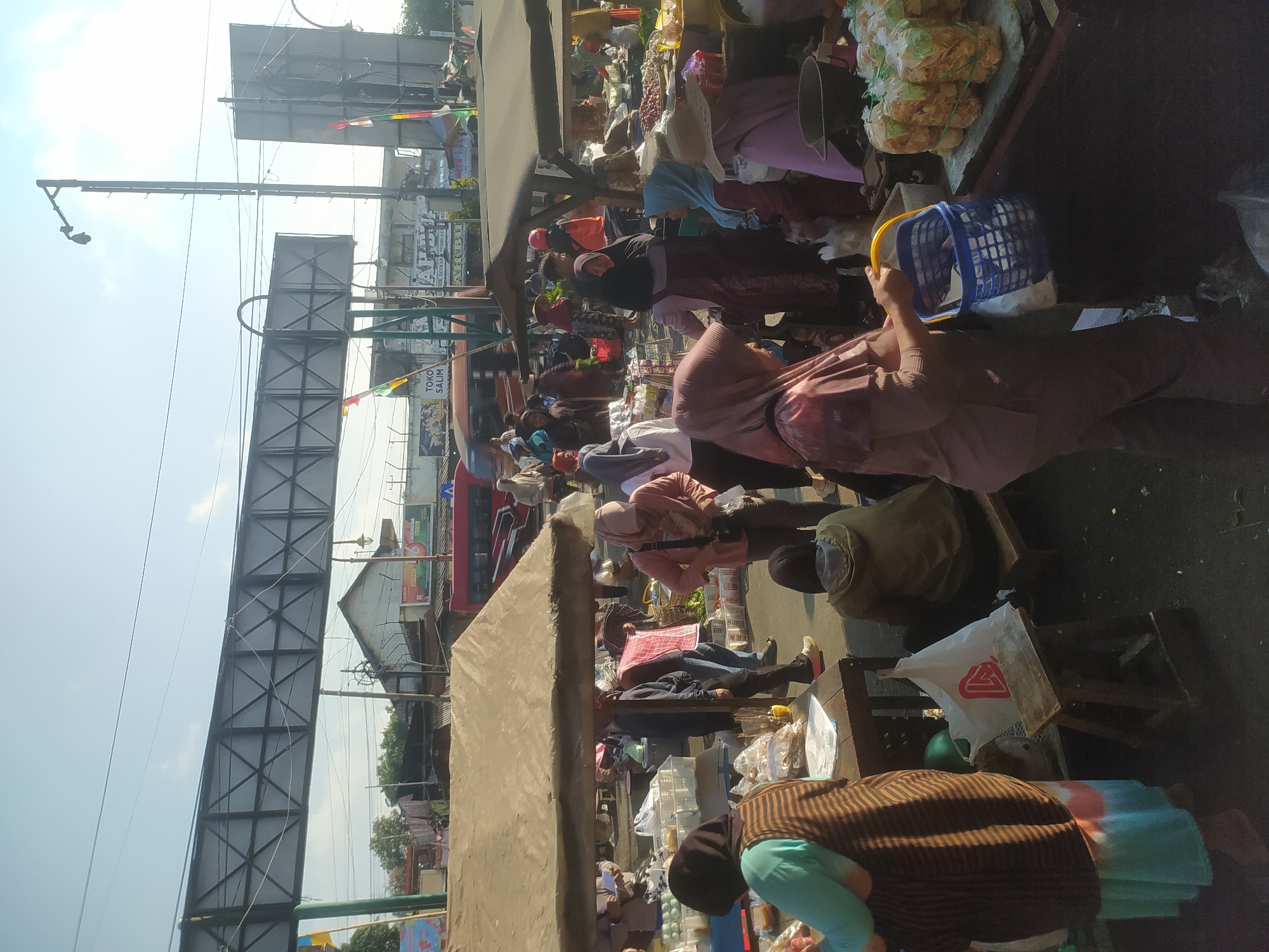 This is a street-level, eye-level, medium shot of a busy outdoor market in Kupang, Indonesia. The scene is filled with people, mostly women, who are shopping and selling various goods. They are dressed in traditional Indonesian attire, with many wearing hijabs. The market stalls are covered with tarps and awnings, providing shade from the sun. Goods are displayed in baskets and on tables, including fruits, vegetables, and packaged snacks. In the background, there are buildings, a red bus, and a large overhead structure, possibly a billboard or a bridge. The overall atmosphere is lively and bustling, suggesting a typical day at the market.