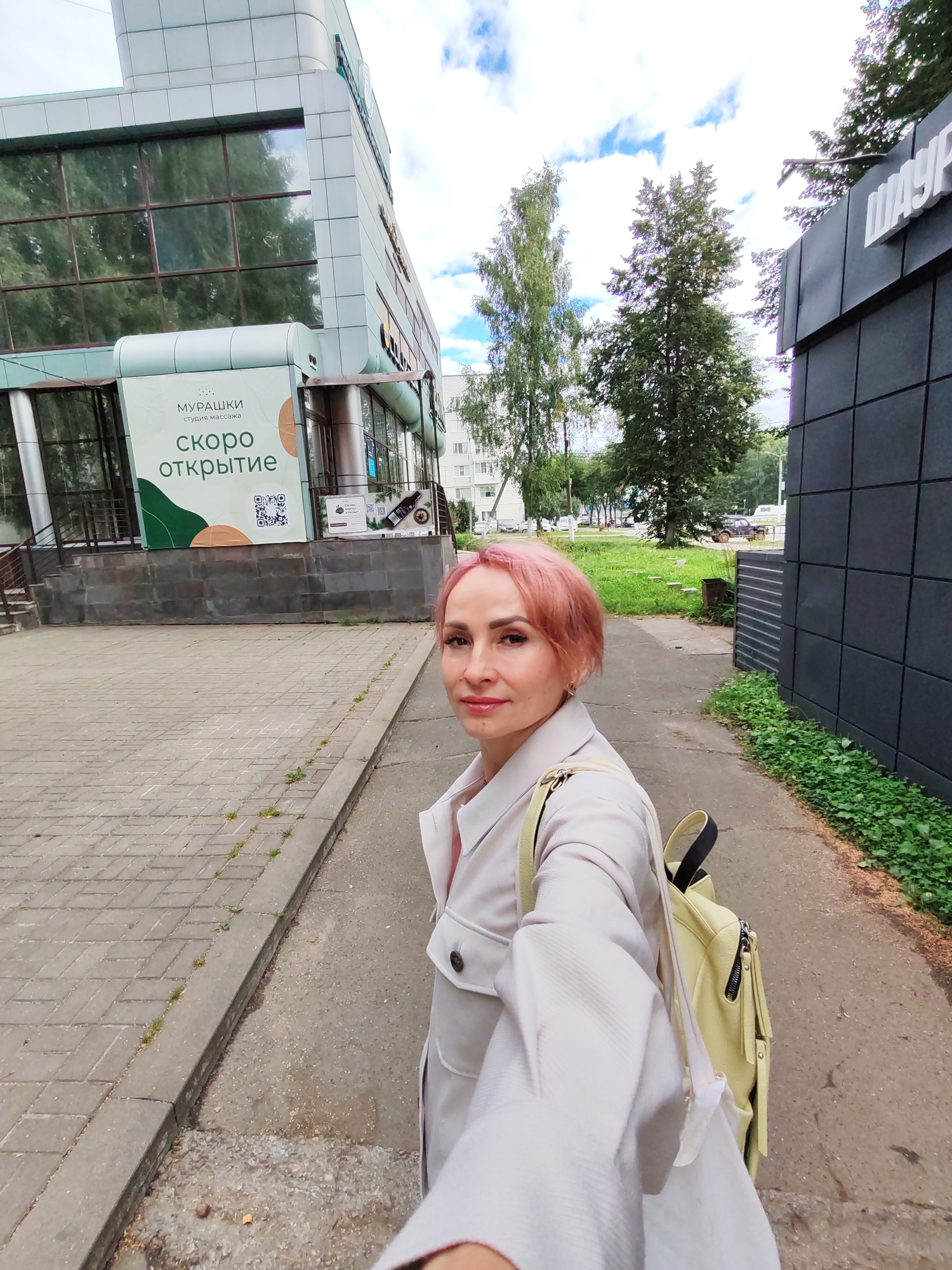 A woman with pink hair is captured in a selfie taken on a sidewalk in Chaikovsky, Russia. She is wearing a light-colored trench coat and a yellow backpack. Behind her, a modern building with large windows and a sign indicating "МУРАШКИ студия массажа скоро открытие" (Murashki Massage Studio Coming Soon) is visible. To the right, a dark-tiled building with white lettering that appears to spell out "ШАУРМ..." (Shaurma...) is present. Trees and a cloudy sky complete the urban landscape.