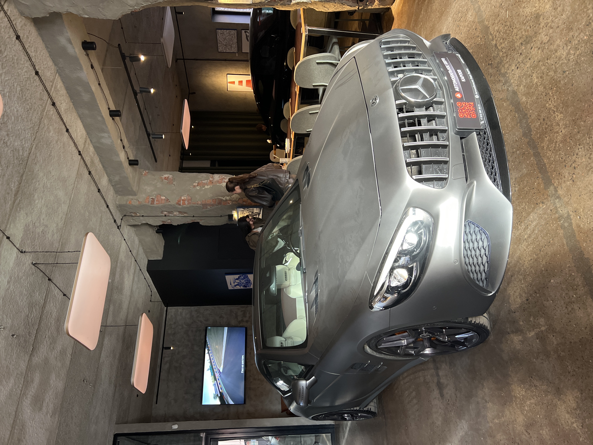 The image captures the interior of a building, possibly a restaurant or an event space, in Saint Petersburg, Russia, as suggested by the text "АВТОМОБИЛЬНОЕ БЮРО КАРПОВ" (Automobile Bureau Karpov) and "+7 812 309 30 21" indicating a Russian phone number. The presence of a sign saying, "Автомобильное Боро Карпов" possibly indicates the establishment's name or a sponsored event.
A gray Mercedes-Benz car is prominently displayed inside the venue. A television screen is located in the background, showing a scene from a race track.
Several people are present, sitting or standing in the background, suggesting they are patrons or staff of the establishment.
The environment features exposed brick and concrete walls with pendant lights, giving it an industrial-chic aesthetic. The overall atmosphere appears modern and stylish.