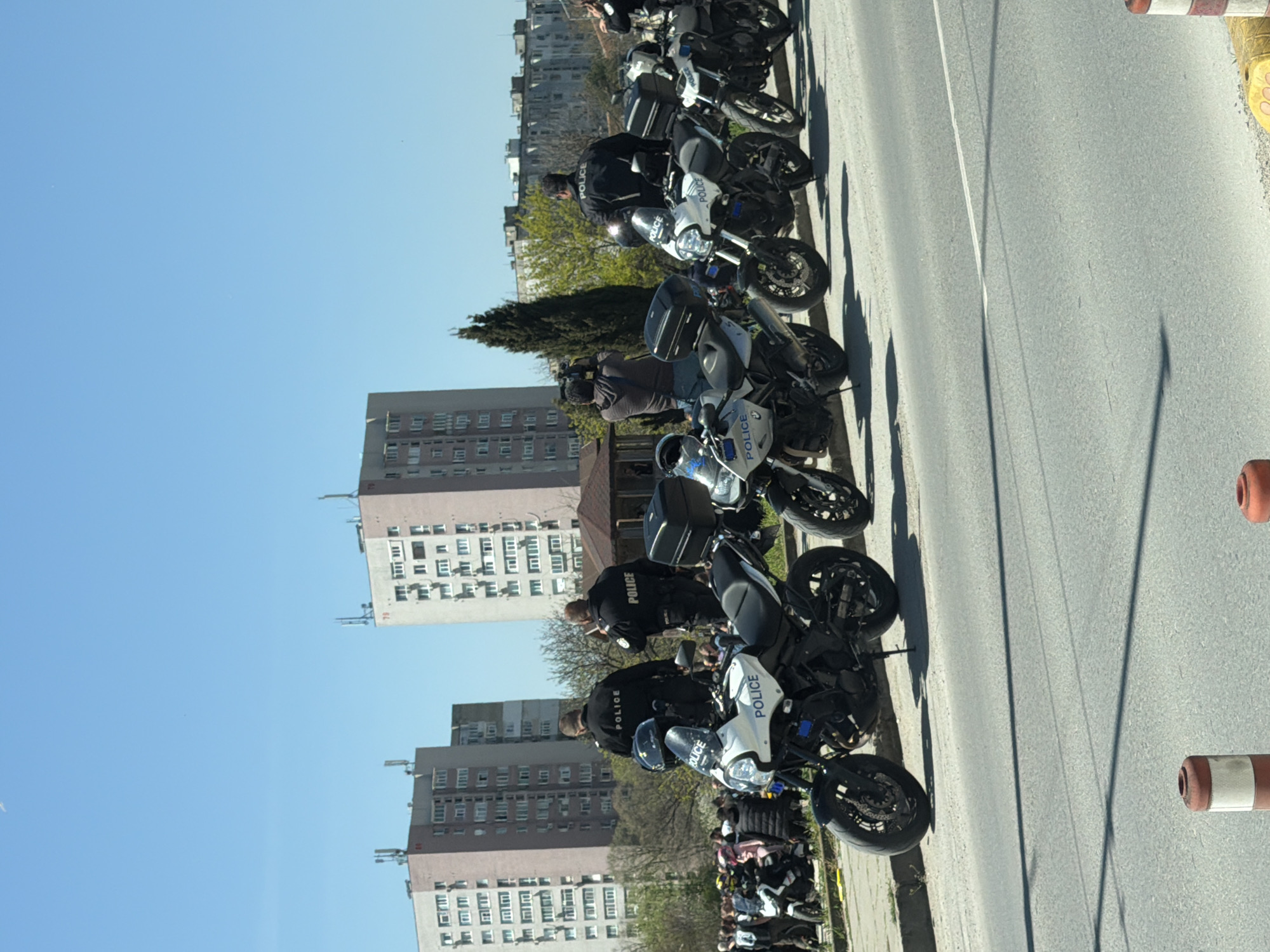 The image captures a scene in Varna, Bulgaria, showing a line of police motorcycles parked on the side of a road, near apartment buildings. The motorcycles appear to be attended by police officers dressed in dark uniforms with the word "POLICE" visible on their backs. The setting is urban, with clear blue skies visible overhead. Orange and white striped traffic cones are also present.