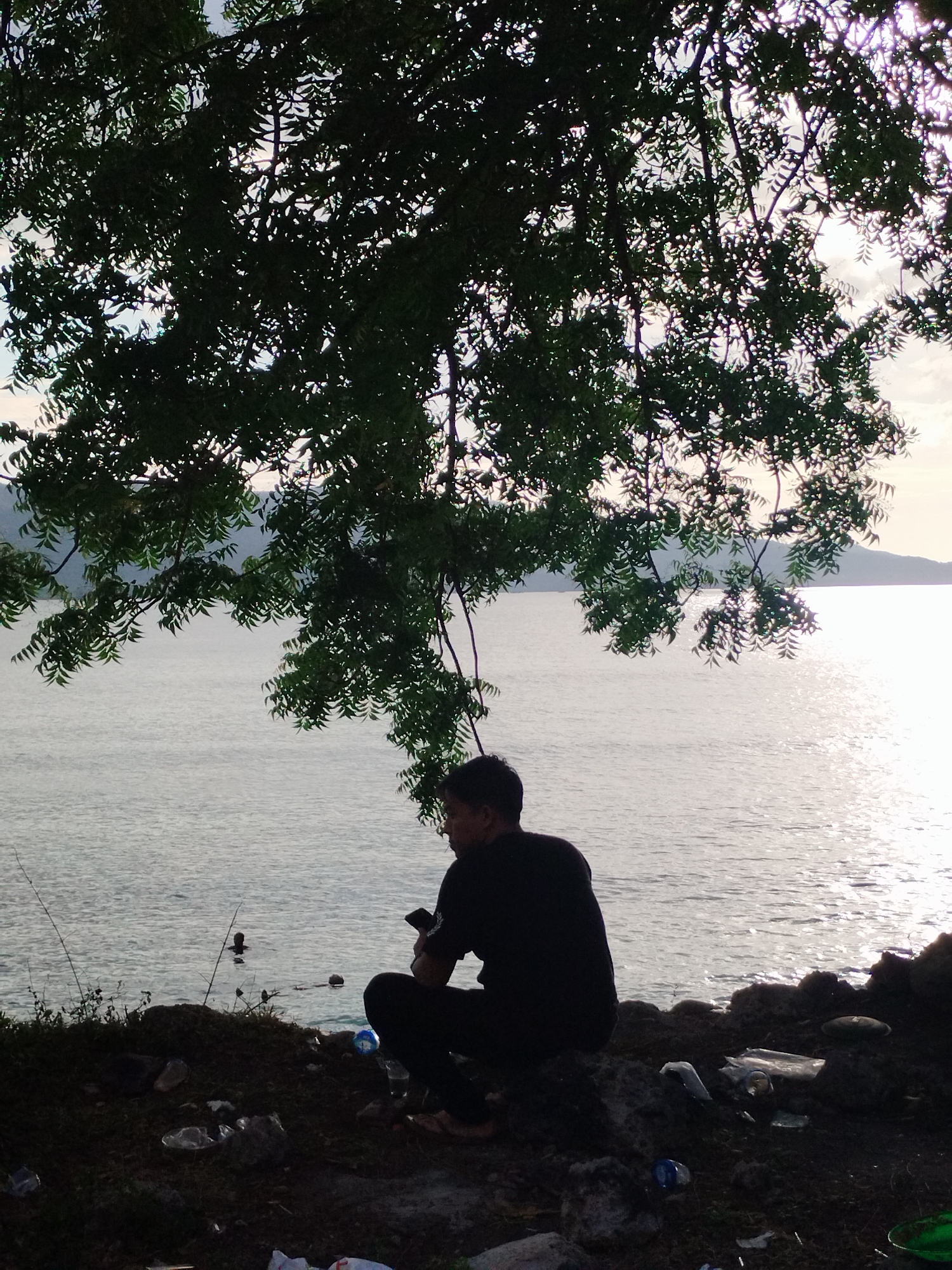 A man is crouched down on the ground by the shore of a body of water, partially obscured by the dark silhouette of a tree hanging overhead. The man is dark-haired and wearing dark clothing, holding a phone in his hand. In the water are two dark shapes, possibly people swimming or objects. The ground near the man is littered with plastic waste. The scene is at Lamreh, Indonesia, judging by the location.