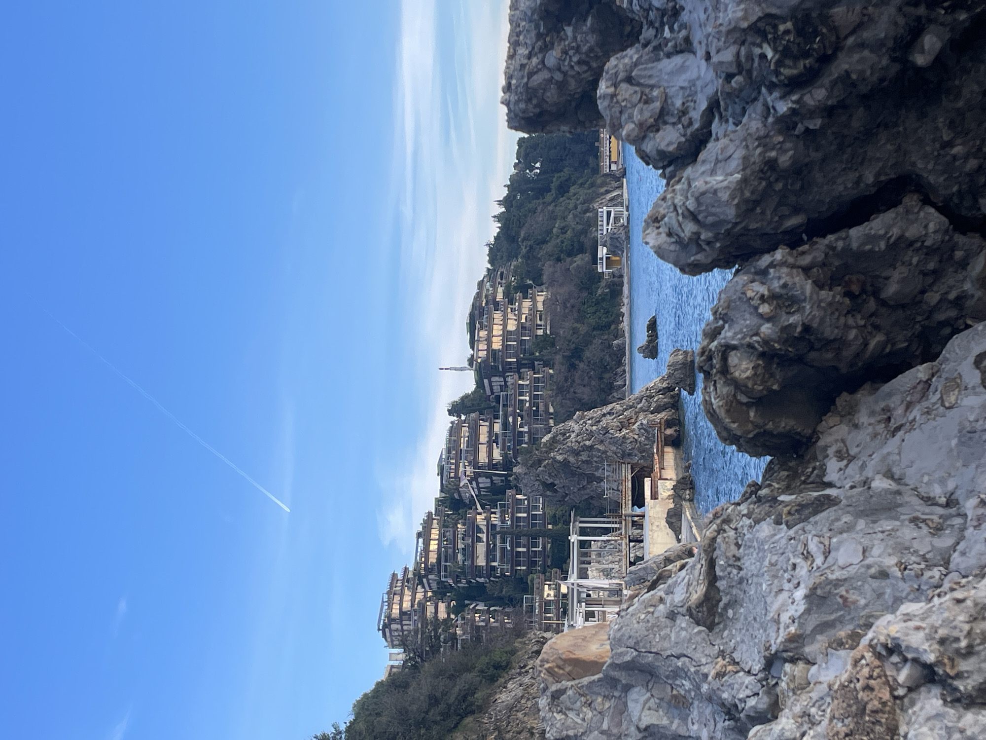 The image displays a coastal landscape in Budva, Montenegro, specifically depicting the Dukley Gardens complex on the Zavala Peninsula. The foreground consists of fragmented, light-colored rocky outcrops, partially out of focus, framing the scene. The midground features calm, deep blue-green water of the Adriatic Sea, with several smaller rock formations protruding from its surface.
Along the immediate coastline, below the main hillside, a white, open-sided structure, possibly a pier or restaurant, is visible on a platform extending into the water, situated beside a prominent, jagged rock outcrop. Further along the coast to the right, a building with yellow and white sections stands at the water's edge.
The background is dominated by a steep, verdant hillside densely covered with trees and shrubs. Integrated into this hillside are numerous multi-story, terraced buildings of the Dukley Gardens complex, characterized by dark architectural frames and reflective glass balconies. The sky above is a clear, bright blue, traversed by a single white aircraft contrail extending diagonally from the upper right. No individuals or specific actions are discernible within the frame. The lighting indicates clear daytime conditions.