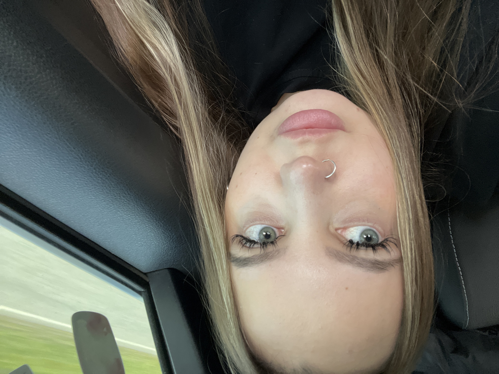 A medium shot shows a Caucasian woman with blonde and light brown hair. She has light blue/grey eyes and a silver nose ring. She appears to be inside a vehicle, possibly a car, as evidenced by the interior trim and a glimpse of the outside through the window. The environment visible outside appears to be a road with green vegetation along the side. The location is not possible to be determined to be in the city of Alexandrovka, Russia.