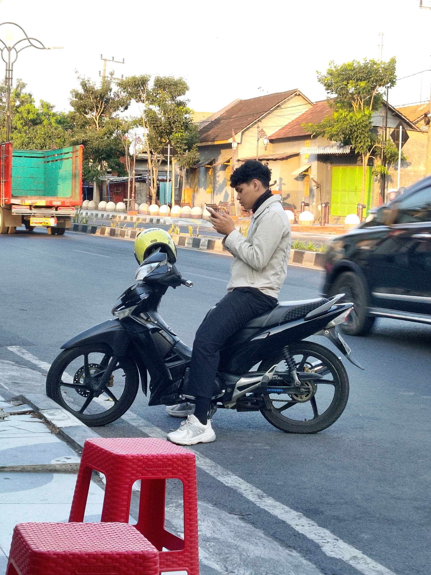 A young man with curly black hair is sitting on a black motorcycle parked on the side of a road in Mojokerto, Indonesia. He is wearing a light-colored jacket, dark pants, and white sneakers, and is looking down at his phone. A yellow helmet rests on the back of the motorcycle. In the background, a green and red truck is visible, along with buildings and trees. The scene appears to be an urban environment during the daytime. Two red plastic stools are placed in the foreground, closer to the viewer. The road has white lane markings, and a concrete curb separates the road from the sidewalk. A black car is partially visible in motion on the road.