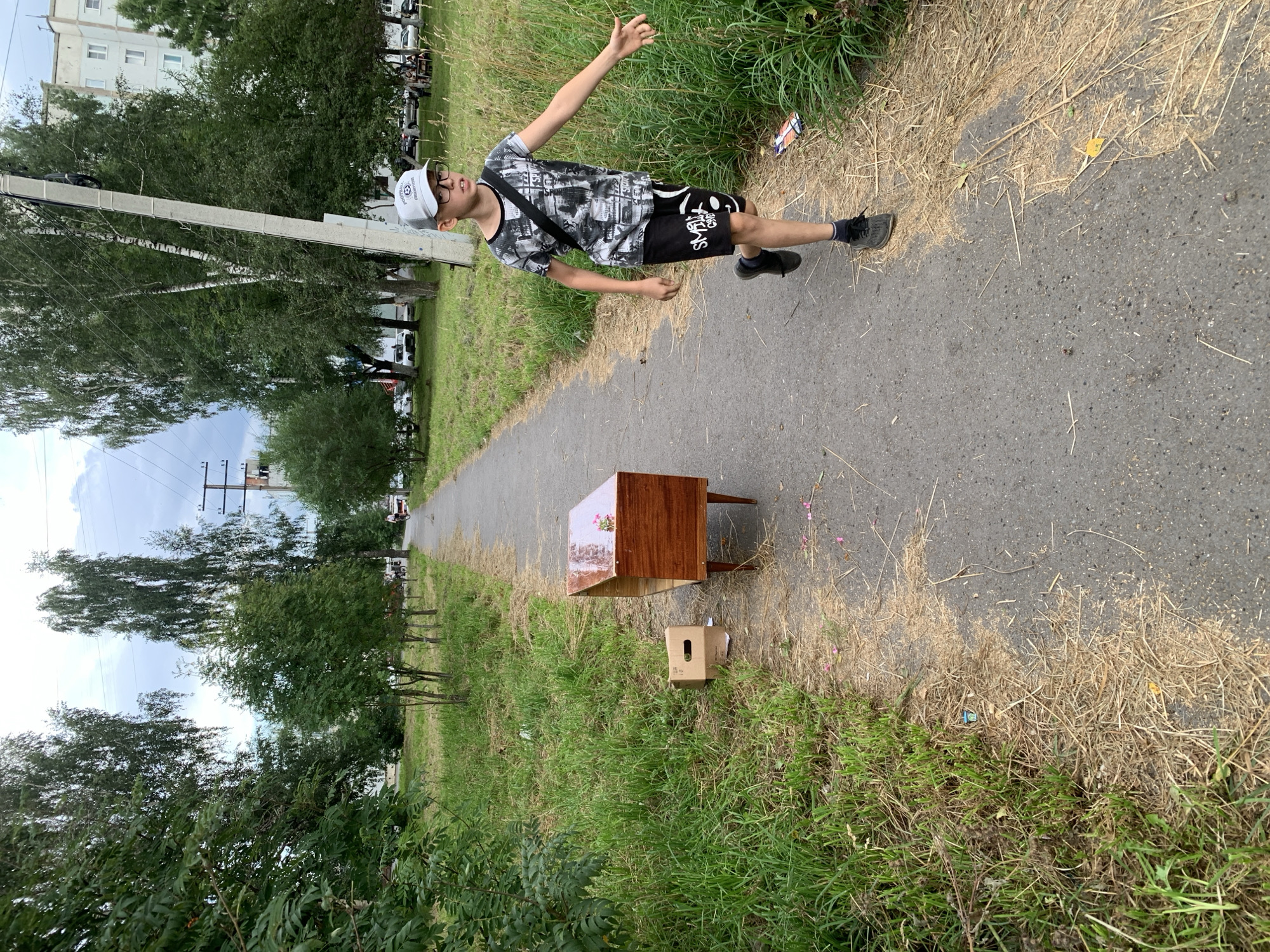 A young male wearing a grey baseball cap, glasses, a t-shirt with a black and white graphic print, and black shorts with a graphic print is walking on a paved path next to a grassy area. He has a black strap across his chest and is wearing dark sneakers. To his left, a wooden piece of furniture, possibly a television stand, sits on the paved path. Beside the furniture is a small cardboard box. The environment appears to be an outdoor urban setting with trees and apartment buildings in the background. The sky is overcast. This scene is located in Prokopyevsk, Russia.