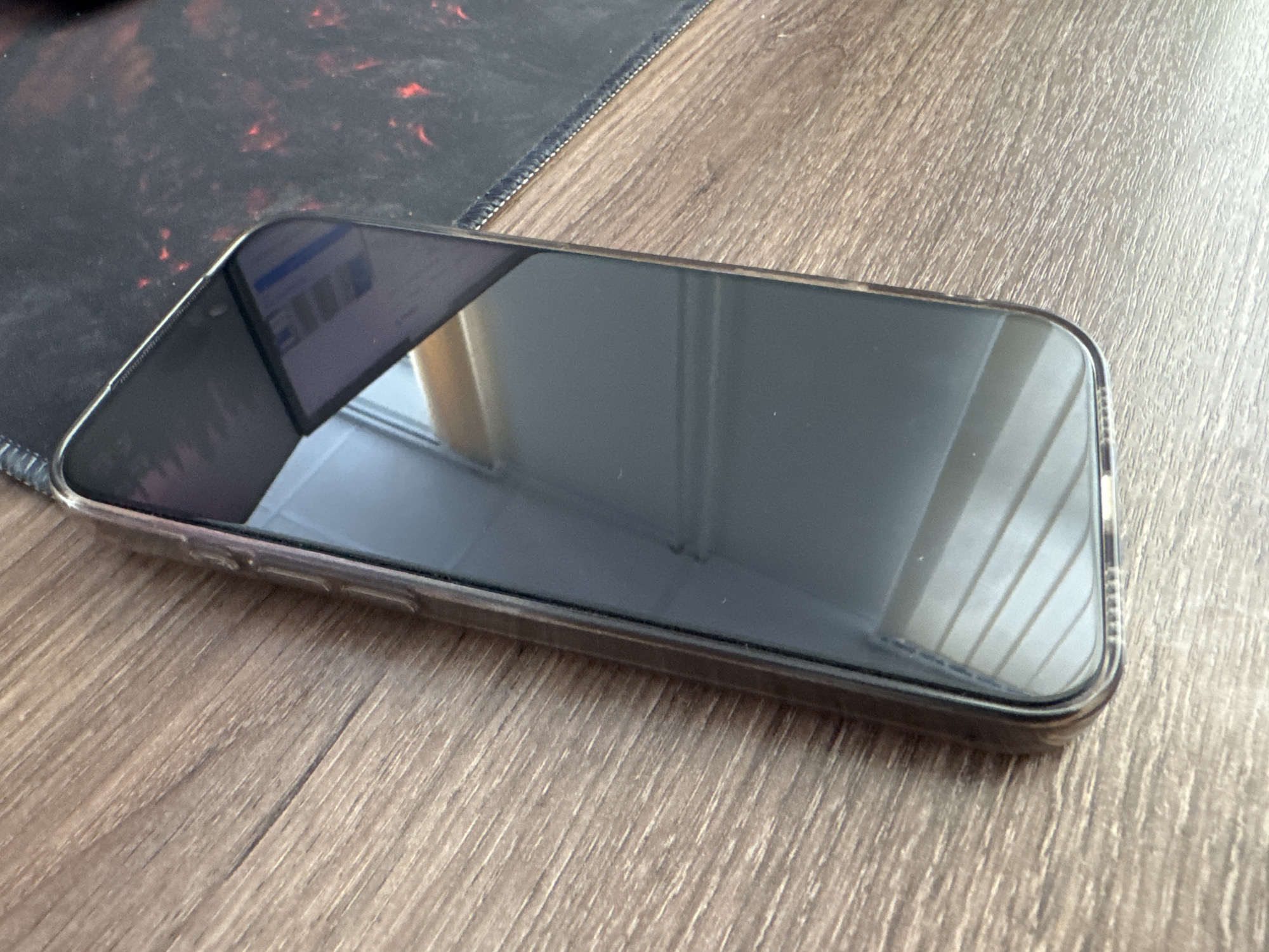 A smartphone in a transparent case is lying on a wooden surface. To the left of the phone, there is a corner of a dark-colored mousepad with a reddish pattern. The phone screen is reflecting the room, suggesting the screen is off or displaying a dark image. The wooden surface appears to be a desk or table. The location is unknown.