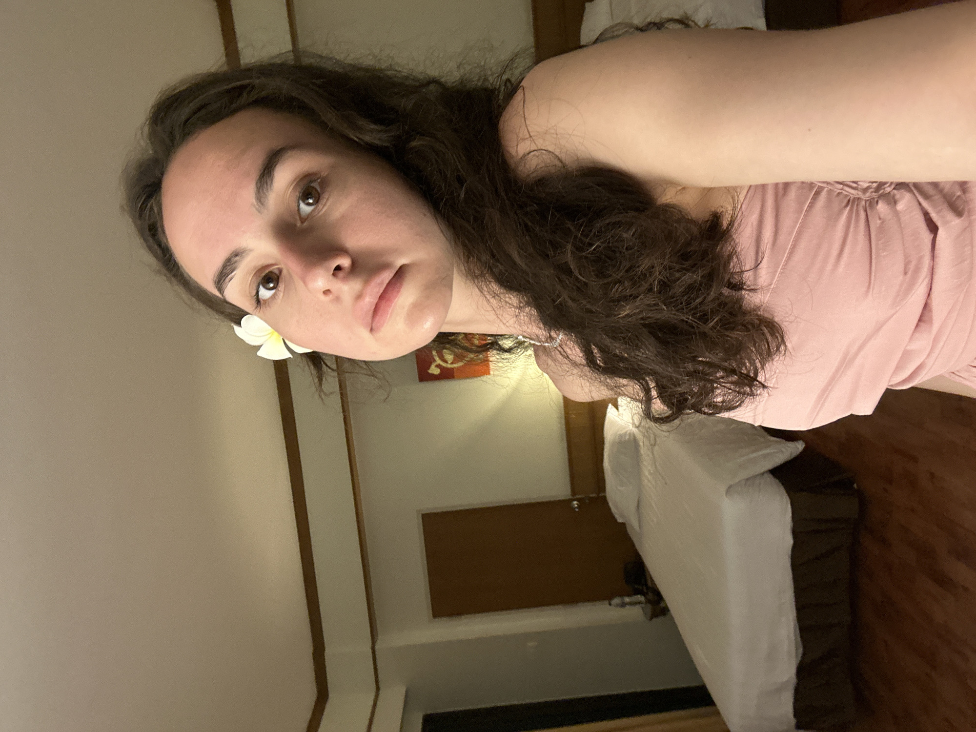 A young woman with dark curly hair is wearing a light pink top and a white flower is tucked behind her left ear. She is indoors in a room with a bed, wooden door, and a small painting on the wall. The room appears to be located in Pattaya City, Thailand.