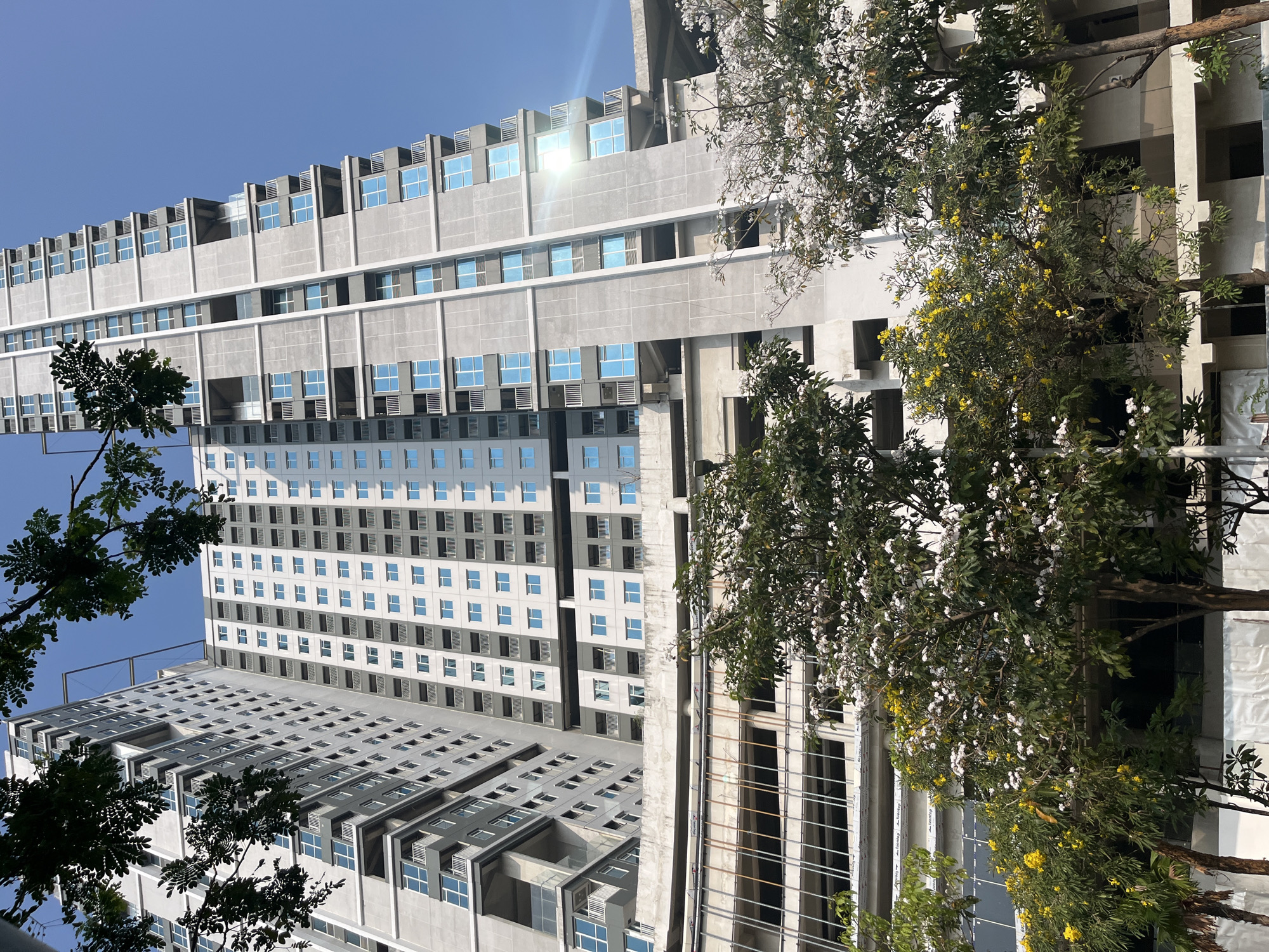 This image captures a contemporary residential building in East Jakarta, Indonesia, under a clear blue sky. The building, a high-rise structure, features a modern architectural design characterized by repetitive rectangular windows, balconies, and a neutral color palette of white and gray. Its facade is composed of segmented sections, with some areas appearing to be under construction or renovation, indicated by exposed metal frameworks and unfinished concrete.
In the foreground, lush green trees with white and yellow blossoms partially obscure the lower levels of the building, suggesting a vibrant natural element contrasting with the urban architecture. The trees are dense, with some branches reaching towards the sky and framing the view of the building. The ground level appears to be a construction site or an area with temporary barriers, with visible support structures and white tarps.
The lighting suggests it is daytime, with sunlight reflecting off the windows of the building and casting subtle shadows. The overall scene depicts a blend of modern urban development and natural elements, characteristic of many rapidly growing cities in Southeast Asia. No people are visible in the frame.