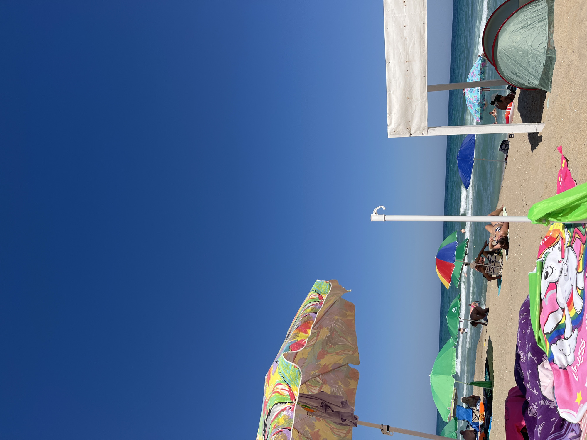 This is an eye-level shot of a crowded beach in Obzor, Bulgaria on a sunny day. The sand is tan, and the ocean is a clear blue with small waves. The sky is a vibrant, cloudless blue. Several beach umbrellas are scattered across the sand, providing shade for beachgoers. In the foreground, a colorful, patterned umbrella is partially visible. Beach towels and a small tent are also spread out on the sand. People are relaxing, sunbathing, and wading in the water. The scene conveys a typical summer beach day with a lively atmosphere.