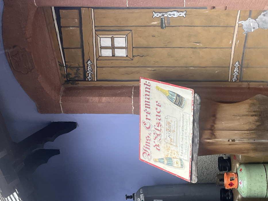 The scene depicts a portion of a building exterior in Kintzheim, France. The left side features a smooth, periwinkle-blue wall with a dark, carved wooden bracket extending from the upper left. To the right, a reddish-brown architrave frames a brown, paneled wooden door. The door is adorned with painted details, including a small, four-pane white window in its upper half, a stylized silver door handle and lock plate, and a silver horizontal decorative bar below the window. Green painted foliage is visible near the top left of the door, and a gray, cartoonish cat figure with white eyes is painted at the door's lower right. In the foreground, positioned centrally, is a weathered rectangular sign with a red border, reading "Vins & Crémant d'Alsace" in ornate script. The sign illustrates two wine bottles, one with a blue label on the left and one with a yellow label on the right. Additional text on the sign references "Hotel & Jean-Michel KOENIG" and "Crée direct du Producteur au Consommateur Cartons de 3.6.12 Bouteilles". This sign rests on a dark, curved support structure. To the immediate left of the sign, partially visible, are several gas cylinders: a tall, light gray cylinder with a black valve at the top and multiple smaller, green cylinders with orange caps in the lower foreground. No people are present in the image.