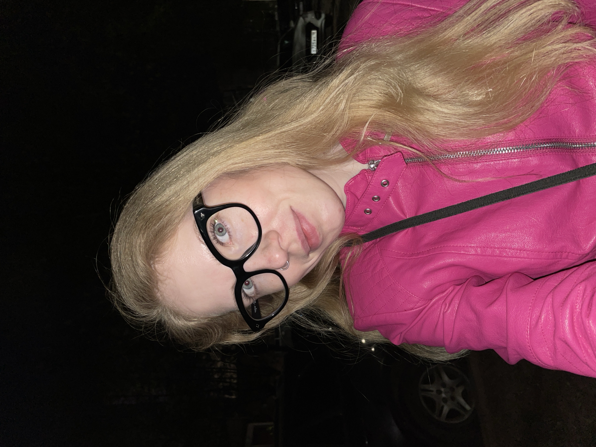 A young adult female is depicted outdoors at night. She is wearing a bright pink leather jacket and black-framed eyeglasses. She also has a nose ring. Her long blond hair is loose and flows past her shoulders. The background is dark, but the wheel of a car is visible in the lower right corner. There is no information in the image that indicates the scene is located in the city of Izhevsk, Russia.