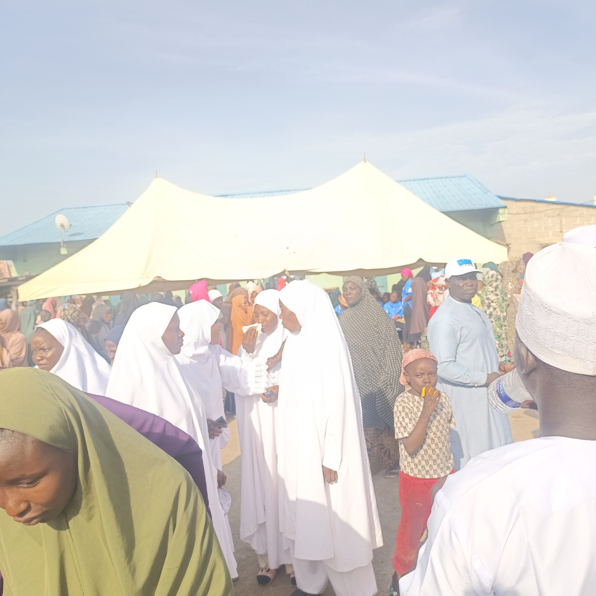 The photograph captures a gathering of people outdoors in what appears to be Akwanga, Nigeria. A large white tent is erected in the background, partially obscuring a blue-roofed building. The foreground is filled with individuals, many wearing traditional Islamic clothing, including white hijabs and robes. Some people are interacting, while others seem to be eating or drinking. A child in a patterned shirt and red pants is eating something. A man in a light blue robe and a white baseball cap stands near the child. The environment appears to be a dusty open area.