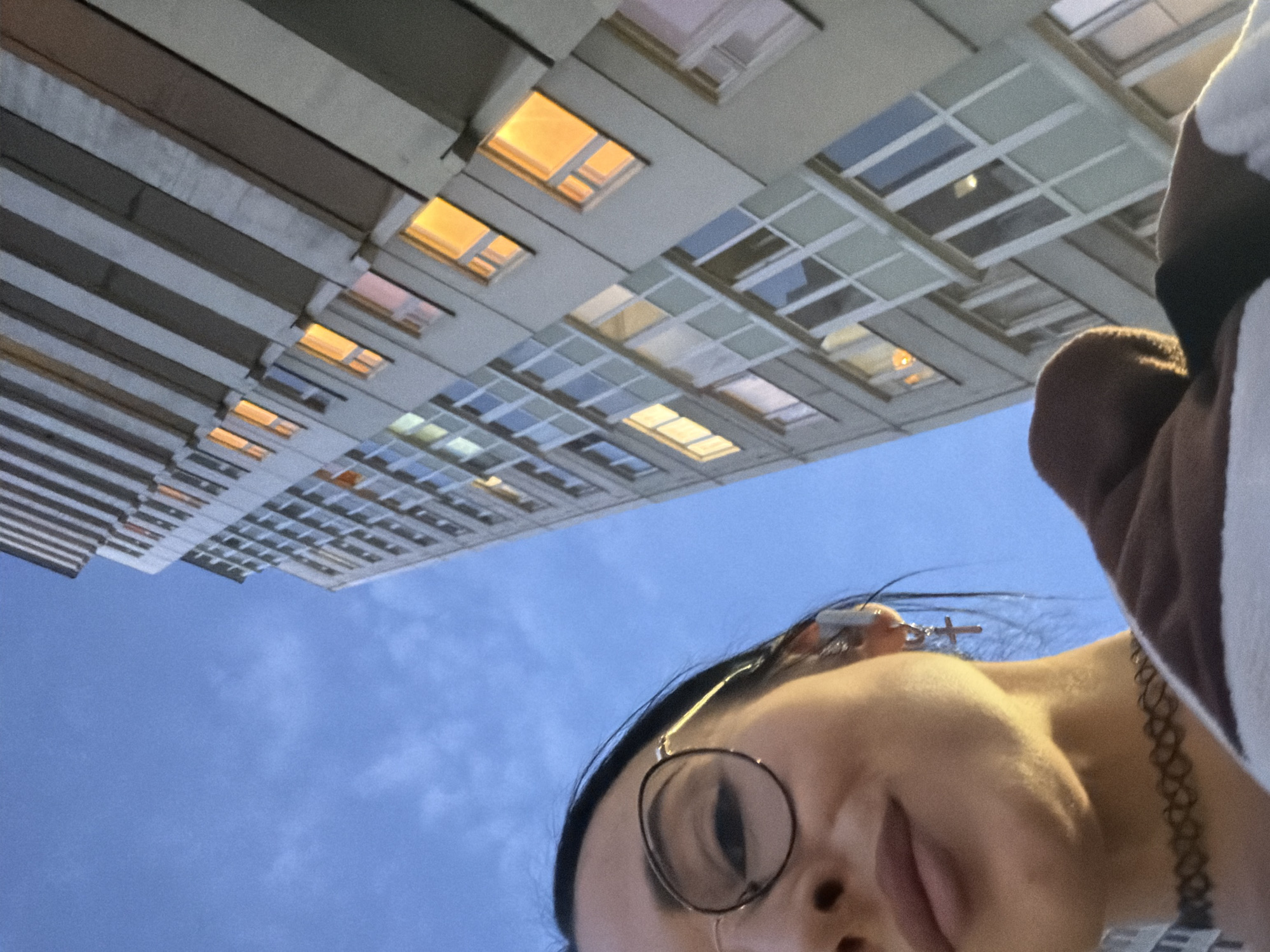 The image is a low-angle selfie, predominantly featuring a person's face in the foreground, looking upwards towards a multi-story urban building against a blue sky.
In the bottom right, the lower portion of a person's face is visible, including their nose, mouth, and chin. They are wearing round-framed glasses, and a silver earring shaped like a cross is visible on their right earlobe. A black, patterned choker necklace adorns their neck, and part of a brown and white garment is seen on their right shoulder. The person's expression is neutral, primarily due to the partial view.
The background is dominated by a modern-style building, which stretches across the top and left side of the frame. The building has a structured facade with numerous rectangular windows, many of which are illuminated with a warm, golden-yellow light. This illumination, combined with the clear, light blue sky featuring faint clouds, suggests the time of day is either dusk or early evening. The architectural style indicates an urban setting, consistent with Saint Petersburg, Russia, where the photo is located. There is no visible text in the image.