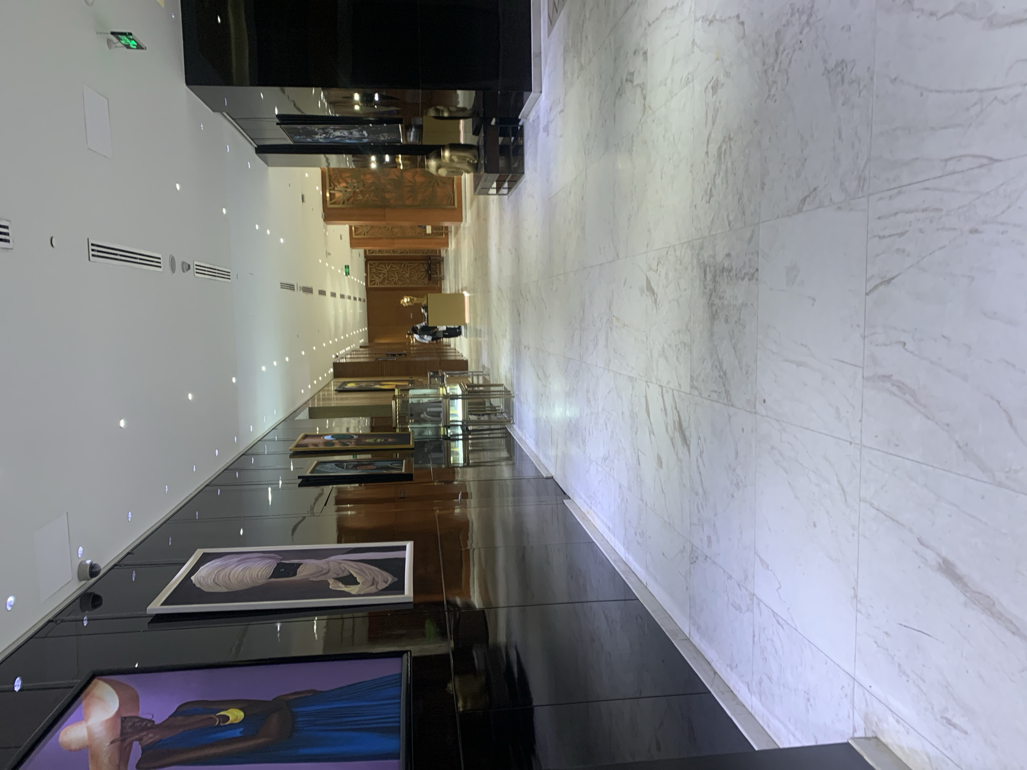 The image shows an interior hallway in a high-end establishment, likely a hotel or gallery in Dar es-Salaam, Tanzania. The hallway features a marble-tiled floor and a wall decorated with framed artwork. The wall is glossy black. The paintings depict figures. The hallway leads to a reception desk with staff present. The lighting is provided by recessed spotlights in the ceiling. There is an emergency exit sign on the left. The ceiling has vents. Security cameras can be seen on the left wall.