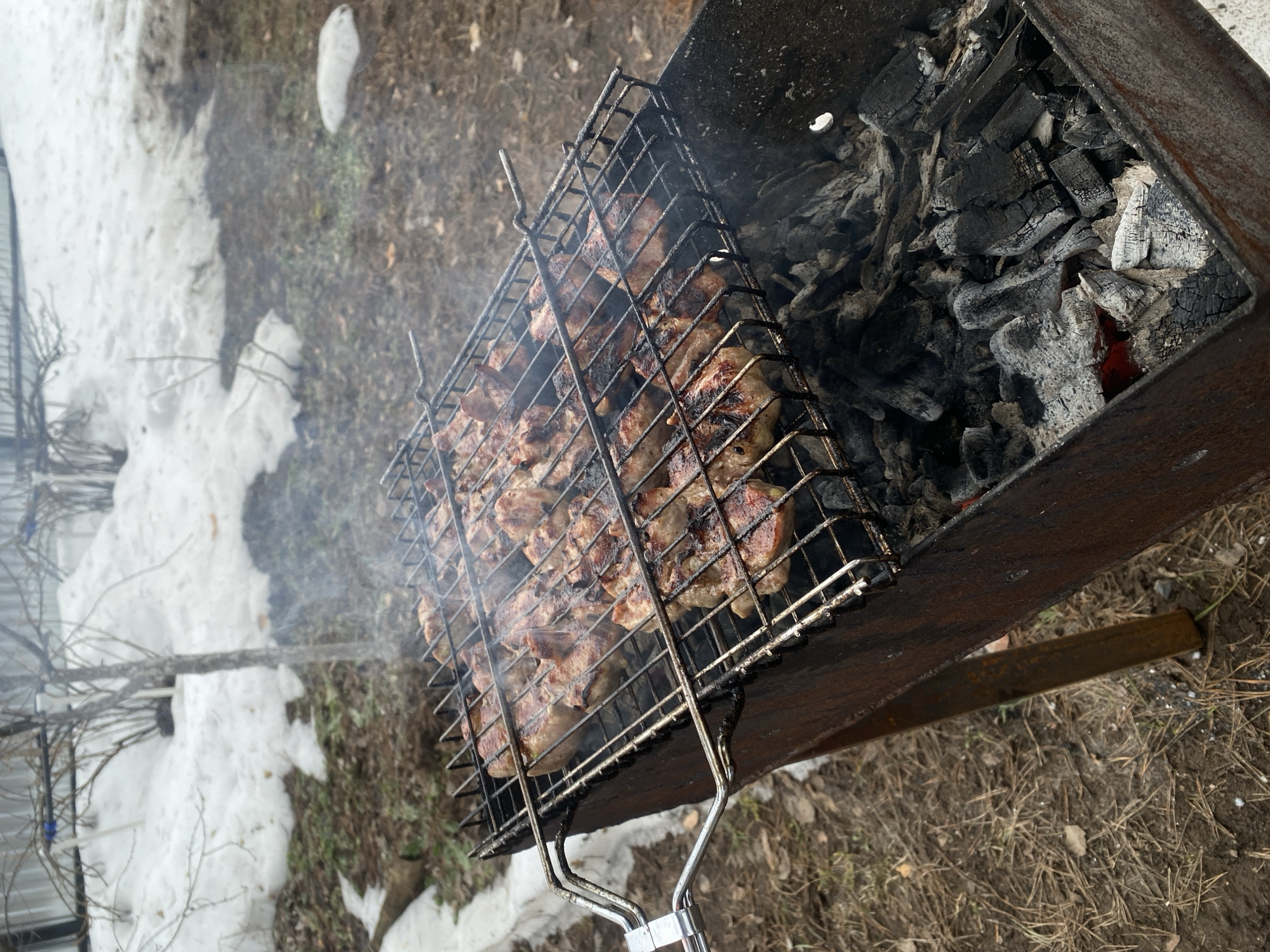 In Chaikovsky, Russia, a metal barbecue grill holds chunks of meat cooking over hot coals. Smoke rises from the grill. Patches of snow remain on the ground, suggesting a cool or early spring day. The area surrounding the grill appears to be a mix of grass and bare earth.