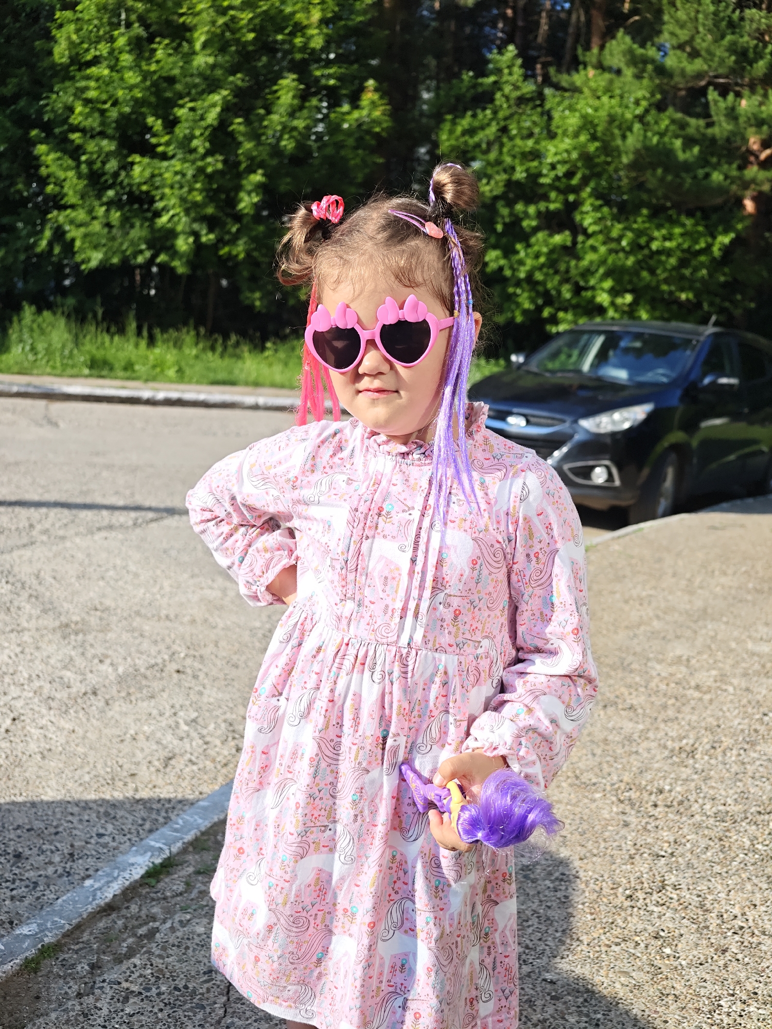 A young girl with purple and pink streaks in her hair, wearing heart-shaped pink sunglasses and a pink unicorn-patterned dress, stands outdoors on a paved surface with a grassy area and trees in the background. A black car is parked in the background. The girl is holding a small purple doll with purple hair. The scene appears to be in a residential or public area in the city of Chaikovsky, Russia.