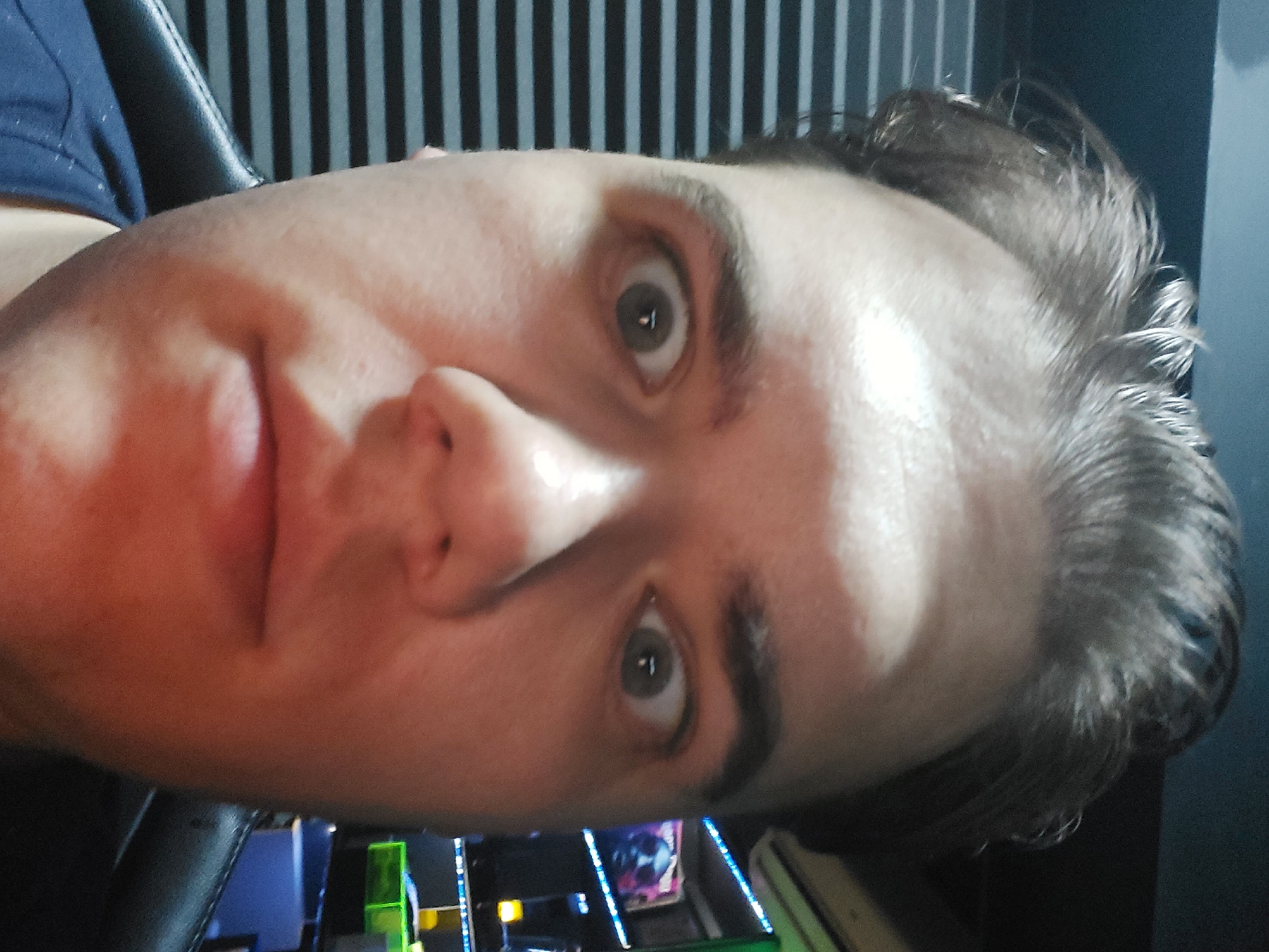 A selfie, taken at a tilted angle, depicts a person with fair skin, brown hair, and gray eyes. The person has a visible nose, lips, and eyebrows. They are wearing a blue shirt. The background features a black and white vertically striped surface and part of a computer keyboard illuminated with blue and green lights. There is no information about the location being Tashkent, Uzbekistan, in the image.