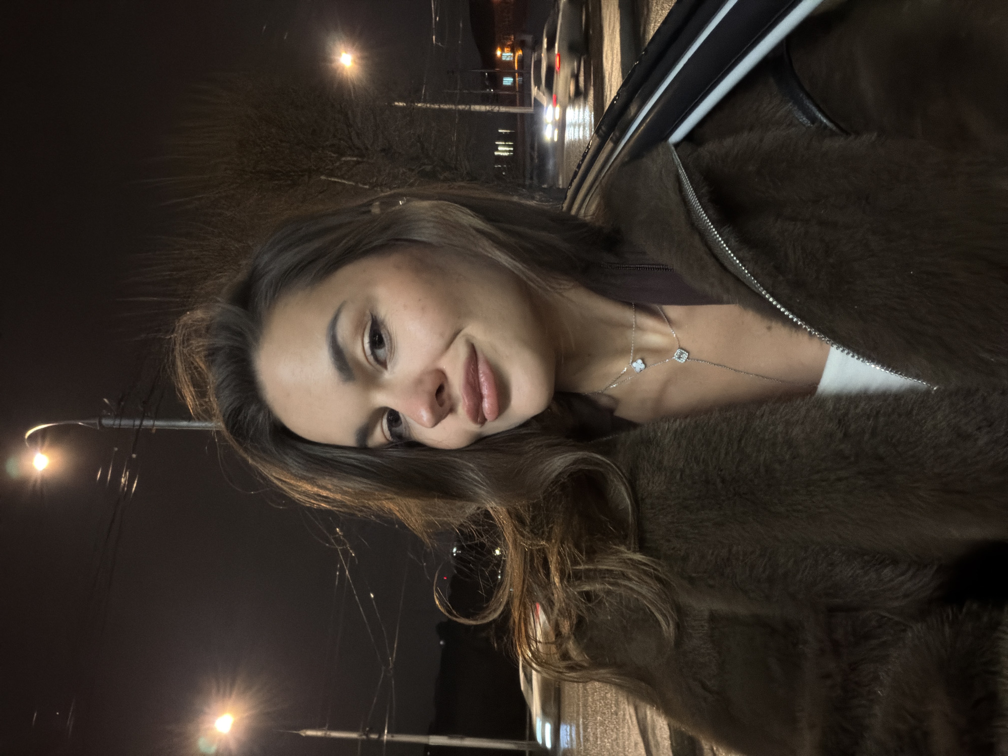 A woman is photographed at night in what appears to be a vehicle in Krasnoyarsk, Russia. The city lights are visible in the background. The woman is wearing a fur coat and a necklace.