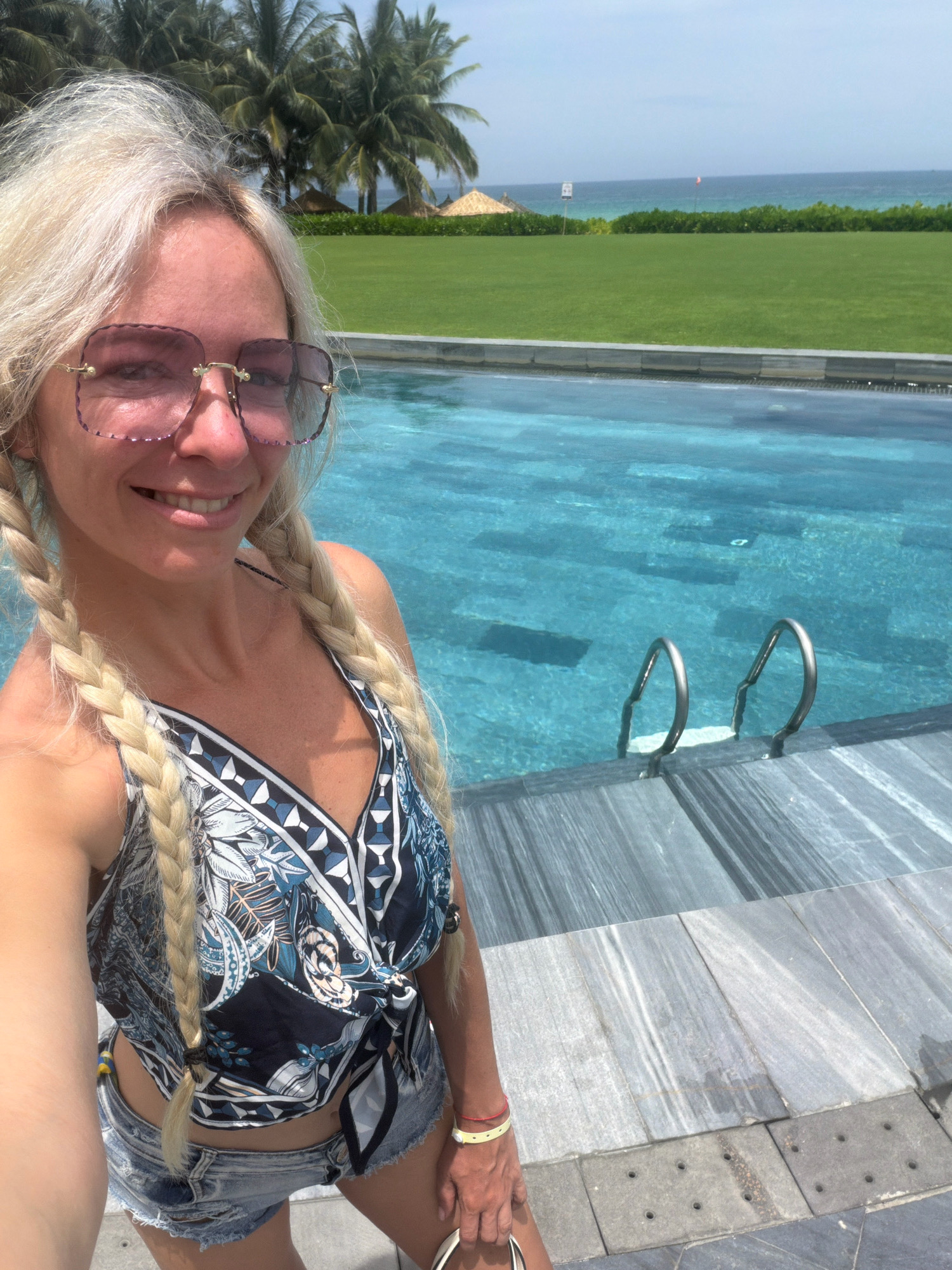 A woman with blonde hair in two braids, wearing oversized pink sunglasses, a blue and white patterned crop top, and denim shorts, poses for a selfie by an infinity pool. The pool has clear blue water and is surrounded by a grey tiled deck. In the background, there is a lush green lawn, palm trees, and the ocean. The scene appears to be a resort in Cam Lâm, Vietnam, likely near a beach.
