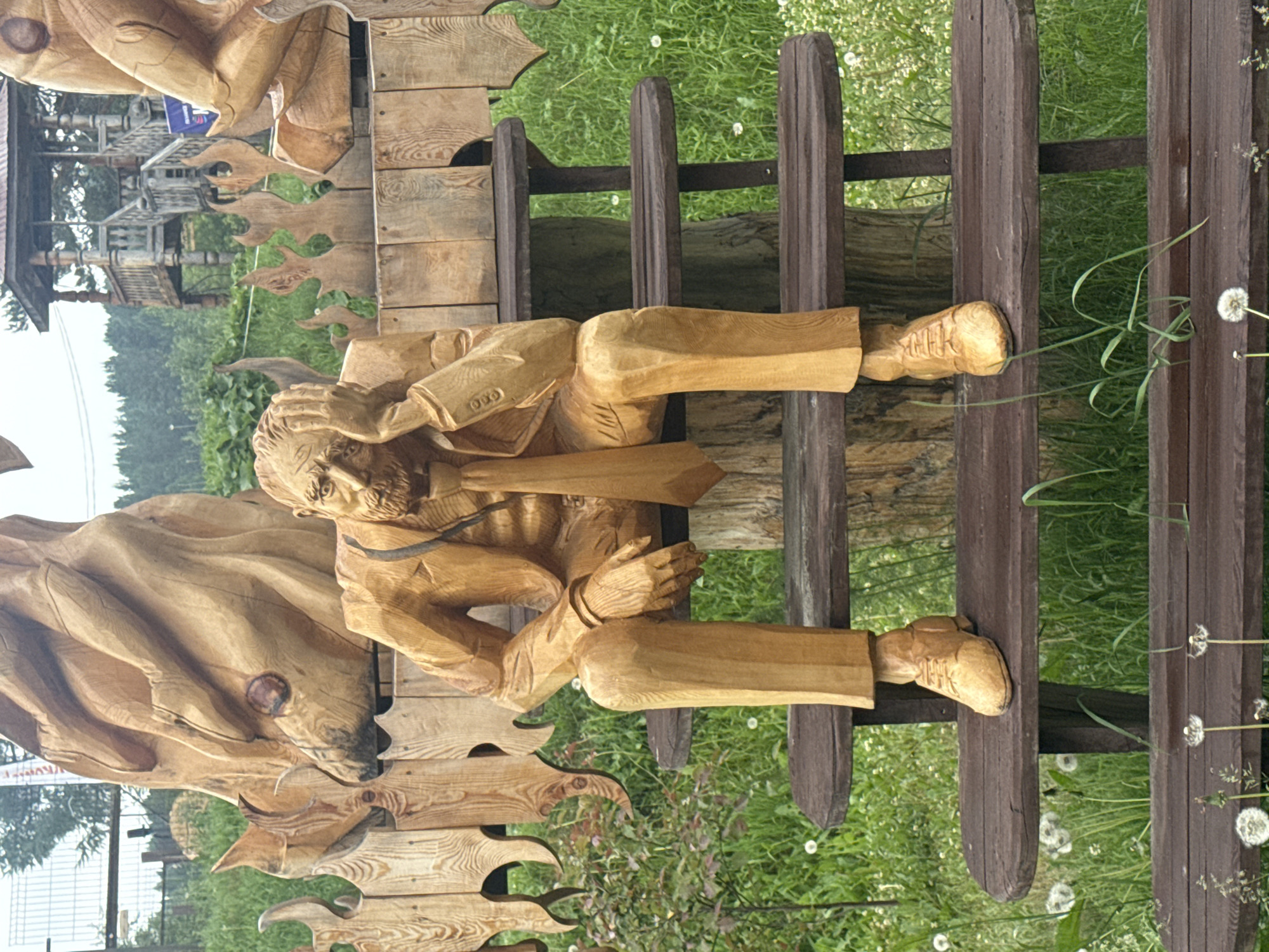 A wooden sculpture of a man in a suit and tie sits on a wooden bench outdoors. The man has his hand on his forehead, appearing distressed. He is positioned in front of a wooden fence with flame-like carvings. In the background, there is a wooden gazebo and trees. The ground is covered in grass and dandelions. The scene is located in the city of Savvateevka, Russia.