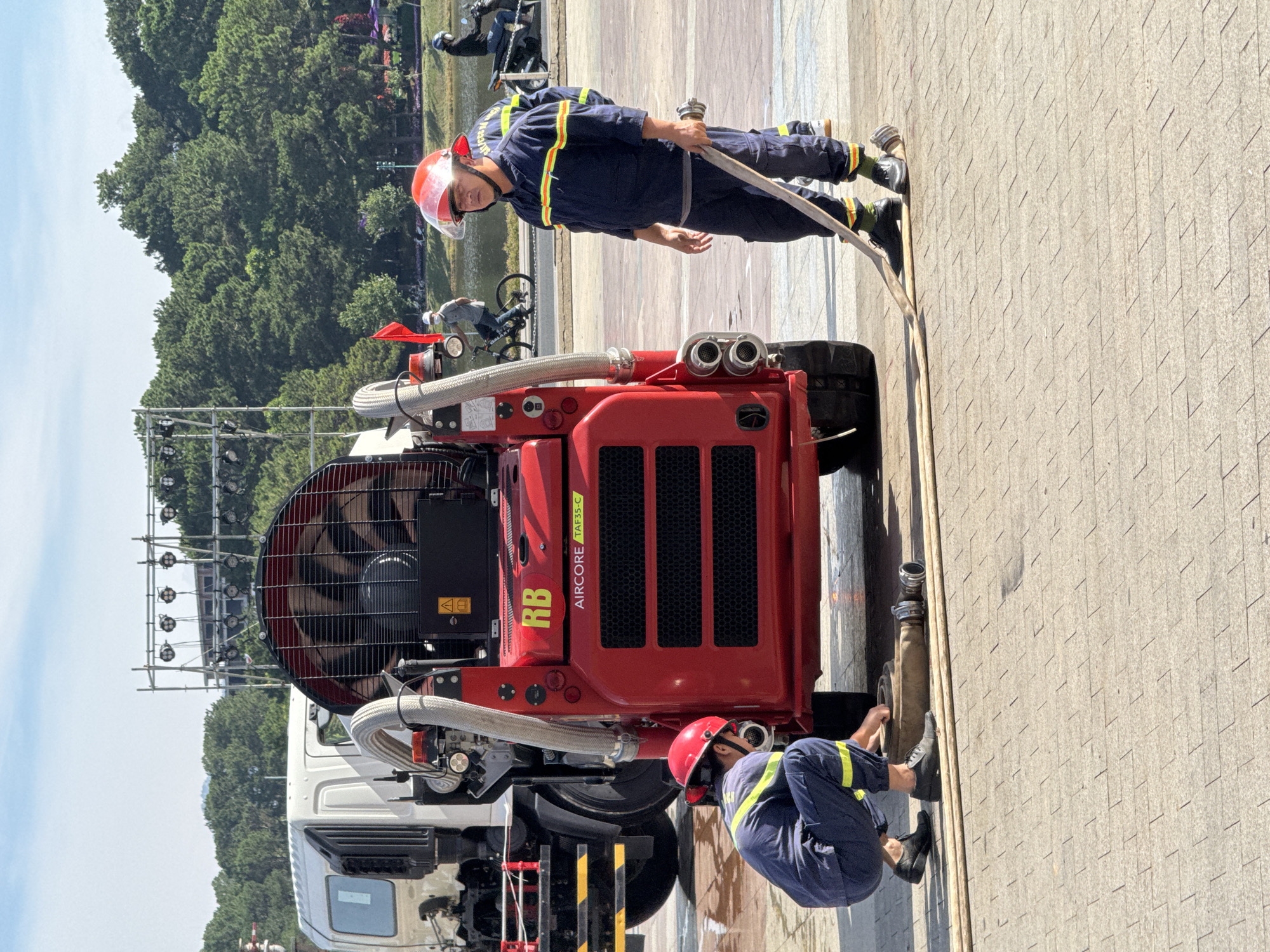 The image captures a scene in Dalat City, Vietnam, near a body of water and a forested area. In the foreground, two individuals in blue uniforms with reflective yellow stripes, presumably firefighters, are interacting with a large red machine, possibly a fire-fighting or water dispersal unit. One individual is crouched down, attending to a hose connected to the machine, while the other stands holding a hose, looking towards the right. The red machine has a large fan at the back and displays the markings "RB" and "AIRCORE TAF35-C". In the background, a white truck is partially visible. Beyond the immediate subjects, a person rides a bicycle near the water's edge, and another person is on a motorcycle. The environment includes paved ground, trees, and a clear sky with some clouds. A stage-like structure with lighting equipment is also visible in the distance.