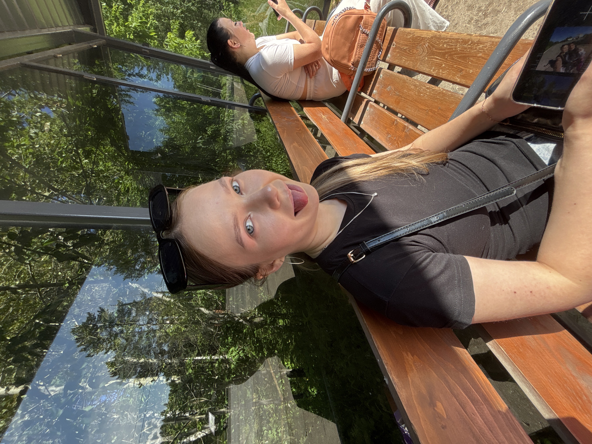 Two young women are sitting on a wooden bench at a bus stop in Chaikovsky, Russia. The woman in the foreground is leaning towards the camera and sticking out her tongue. She has on a black t-shirt and a silver necklace. She is wearing large hoop earrings and her hair is pulled back. The woman in the background is wearing a white t-shirt and light-colored pants. She is looking at her phone and has a brown purse on the bench next to her. The bus stop has large glass windows that reflect the surrounding trees and greenery. The sun is shining, casting shadows on the bench and the ground.