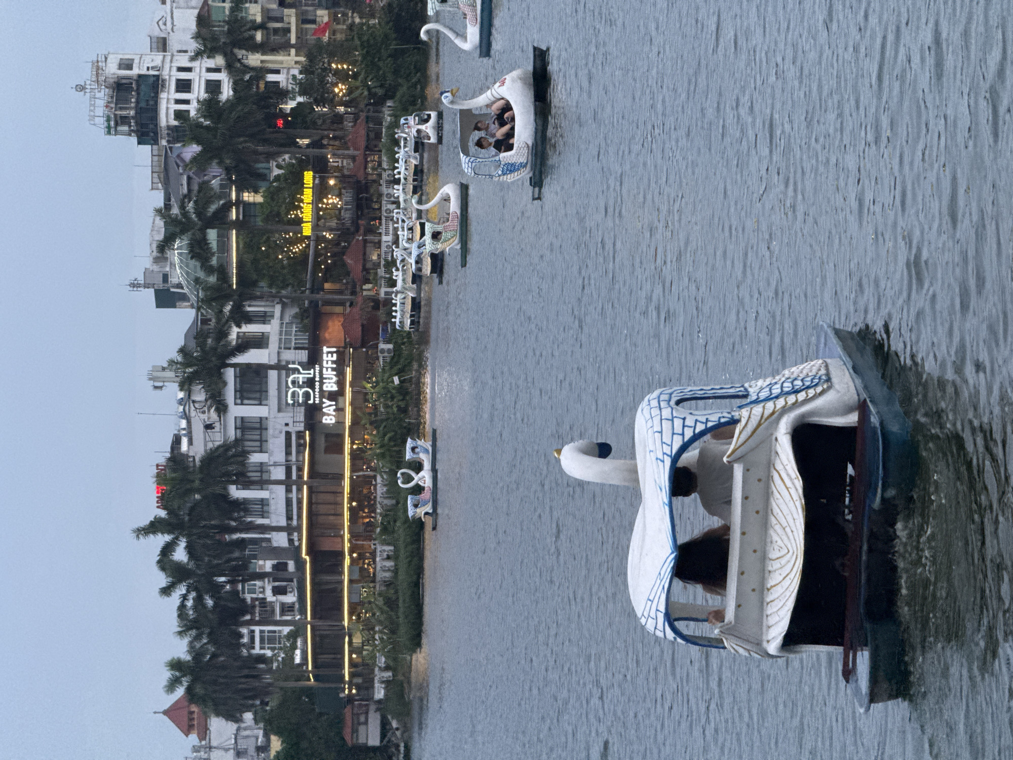 The image depicts a lake with several swan-shaped pedal boats. In the foreground, a white swan pedal boat is moving away from the viewer, with two people seated inside. The boat has a white canopy with blue trim. Further in the background, other swan boats are visible on the water, some with occupants. Across the lake, a long, multi-story building is situated, featuring a prominent sign that reads "BAY BUFFET" with smaller text above it, likely indicating it is a seafood buffet. Palm trees are planted in front of the building, and the cityscape with buildings and a tower is visible behind the trees. The sky is overcast, suggesting it is either early morning or late afternoon. The overall scene appears to be a recreational area in Hanoi, Vietnam, possibly West Lake, given the presence of swan boats and the architecture.