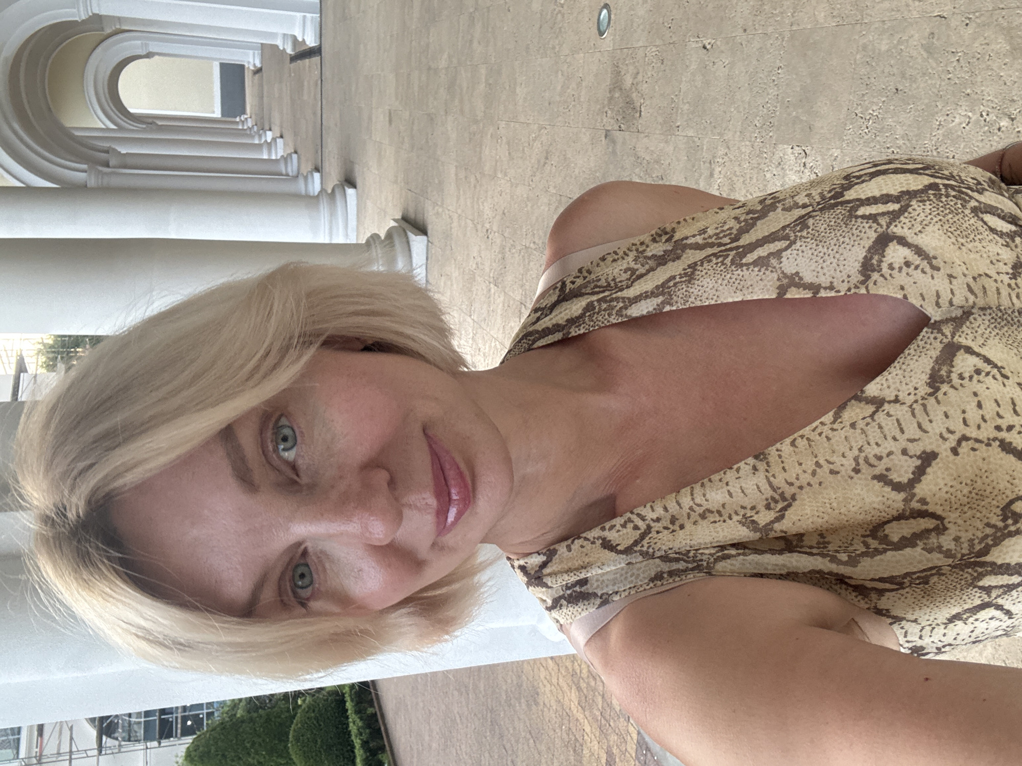 This is a portrait of a woman with blonde hair and blue eyes. She is wearing a light-colored dress with a snake-print pattern. The image is taken outdoors, with a backdrop of white columns and a stone walkway. The lighting suggests it is daytime. The woman's face is turned towards the camera, and she has a slight smile. The background features architectural elements, possibly a colonnade or arcade, with a hint of greenery and a building in the distance. The orientation of the image is slightly tilted.