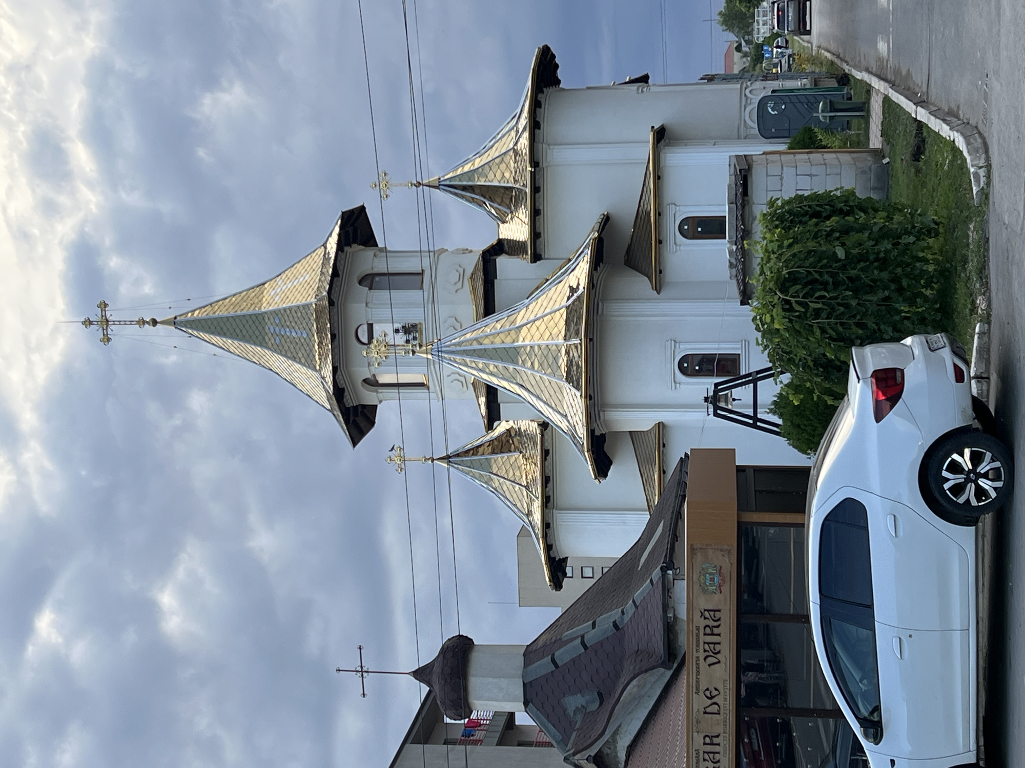 The image captures an exterior view of a white church with multiple golden-domed spires against a cloudy sky. The church has a traditional architectural style, with arched windows and crosses atop the spires. In the foreground, a white sedan is parked on the street. Adjacent to the church is a building with a brown facade and signage that reads "BAR DE VARĂ". The scene is situated in the city of Năvodari, Romania. The street in front of the church is paved, with a sidewalk and some greenery. Power lines are visible across the upper portion of the image. The overall atmosphere is somewhat overcast, suggesting a potentially dull or rainy day.