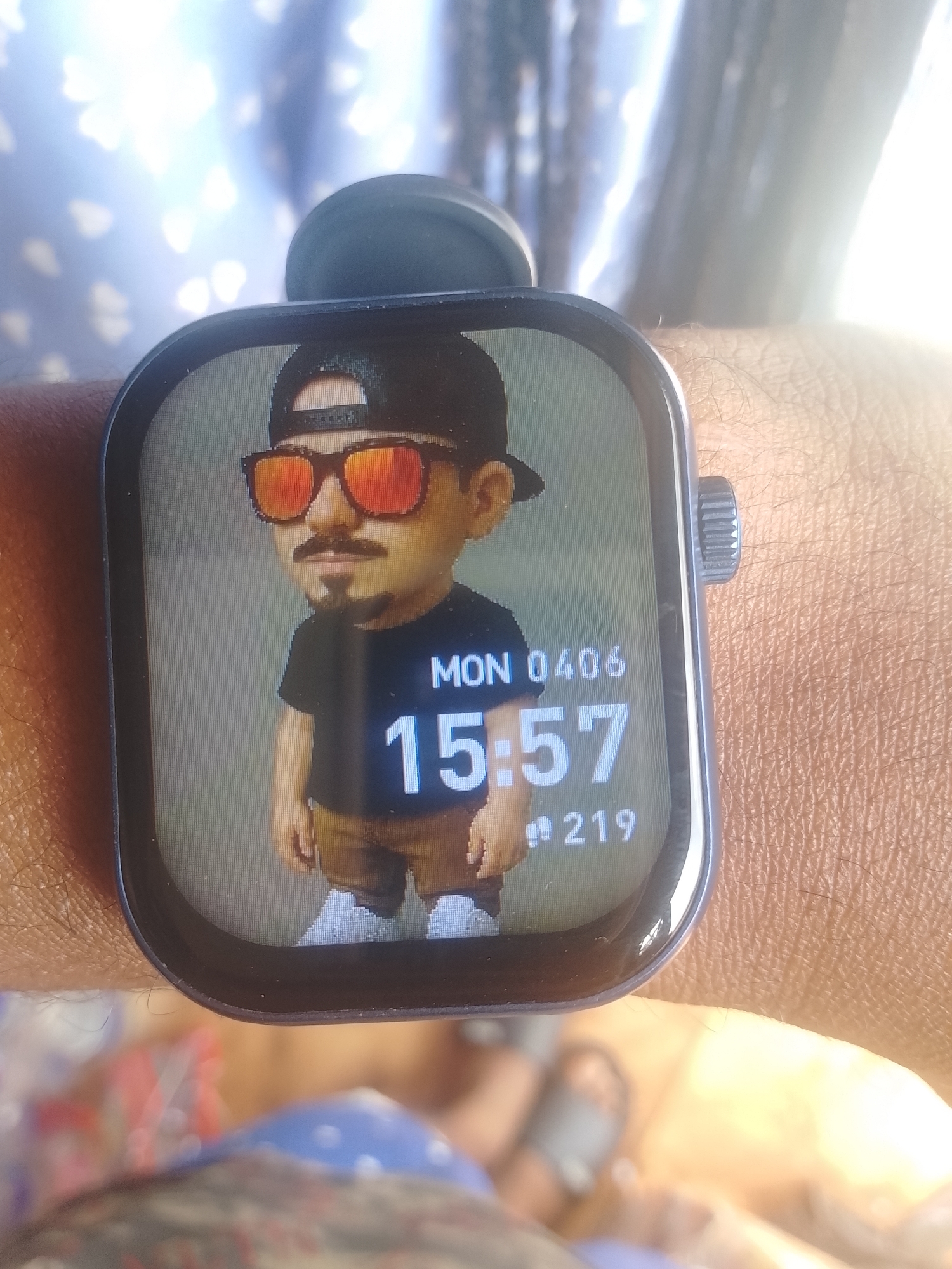 A smartwatch is displayed on a wrist. The watch face shows a cartoon avatar of a man wearing a backwards baseball cap and sunglasses, a black T-shirt, and brown pants. The date and time displayed on the watch are "MON 0406" and "15:57," respectively. The number "219" is also visible at the bottom of the watch face. The arm and wrist on which the watch is worn appear to be a light-to-medium brown skin tone. The background is blurry, but appears to include patterned fabric and possibly a sandal-clad foot. The location is Ejule, Nigeria.