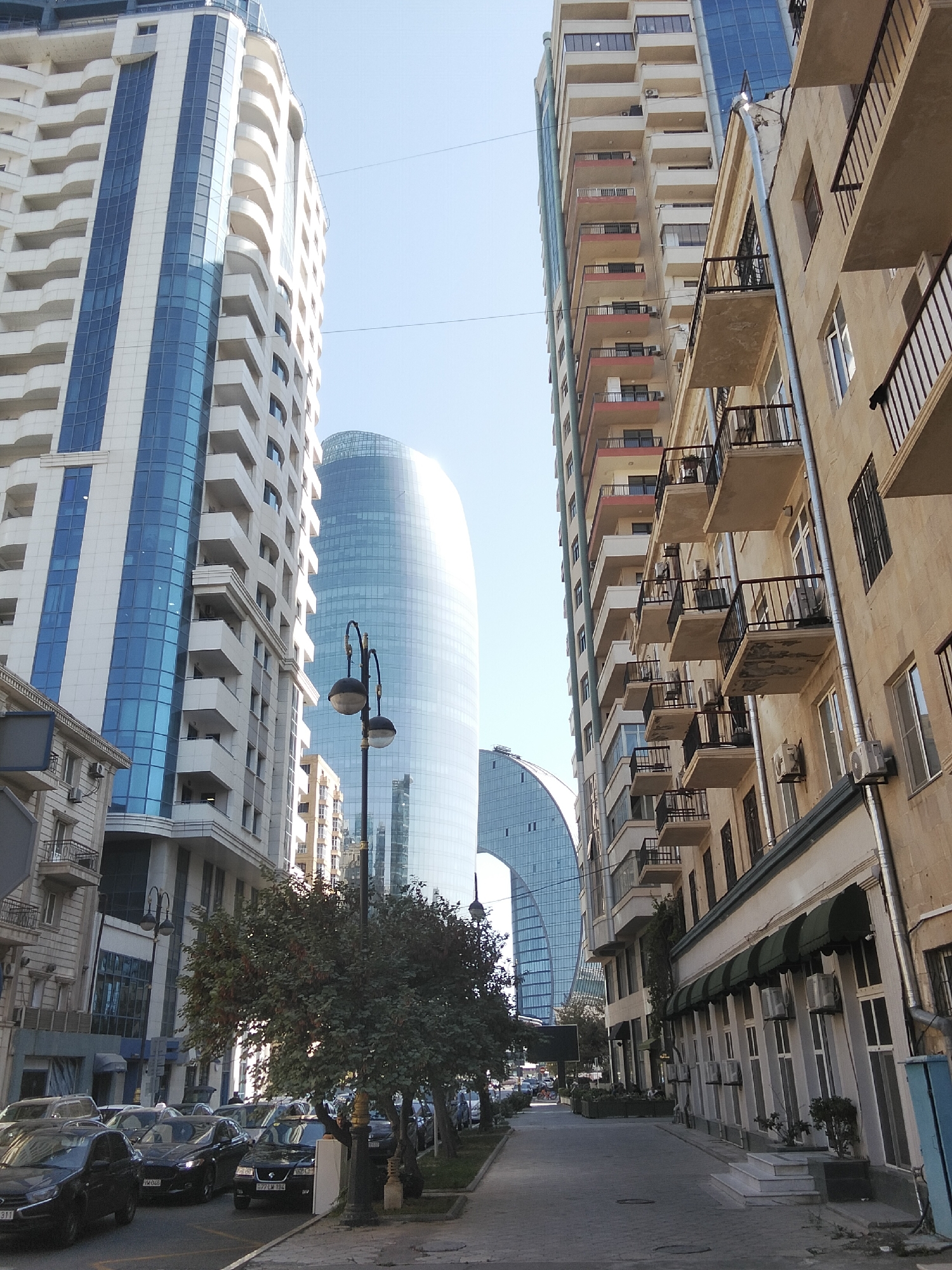In Baku, Azerbaijan, a street scene unfolds with a mix of modern and older architecture. Tall, contemporary buildings with sleek glass facades dominate the background, featuring prominent structures such as the Flame Towers, visible as distinctive curved, glass-clad skyscrapers. In the foreground, a more traditional, beige-colored building with multiple balconies lined with black railings stands alongside a lighter colored, modern apartment building with blue-tinted windows. The street is lined with parked cars, predominantly dark-colored sedans, and a few trees provide greenery along the sidewalk. A lamppost with two spherical lights stands near the trees. The sky is clear and blue, suggesting a bright, sunny day. The scene captures a dynamic urban environment, showcasing the city's architectural evolution.