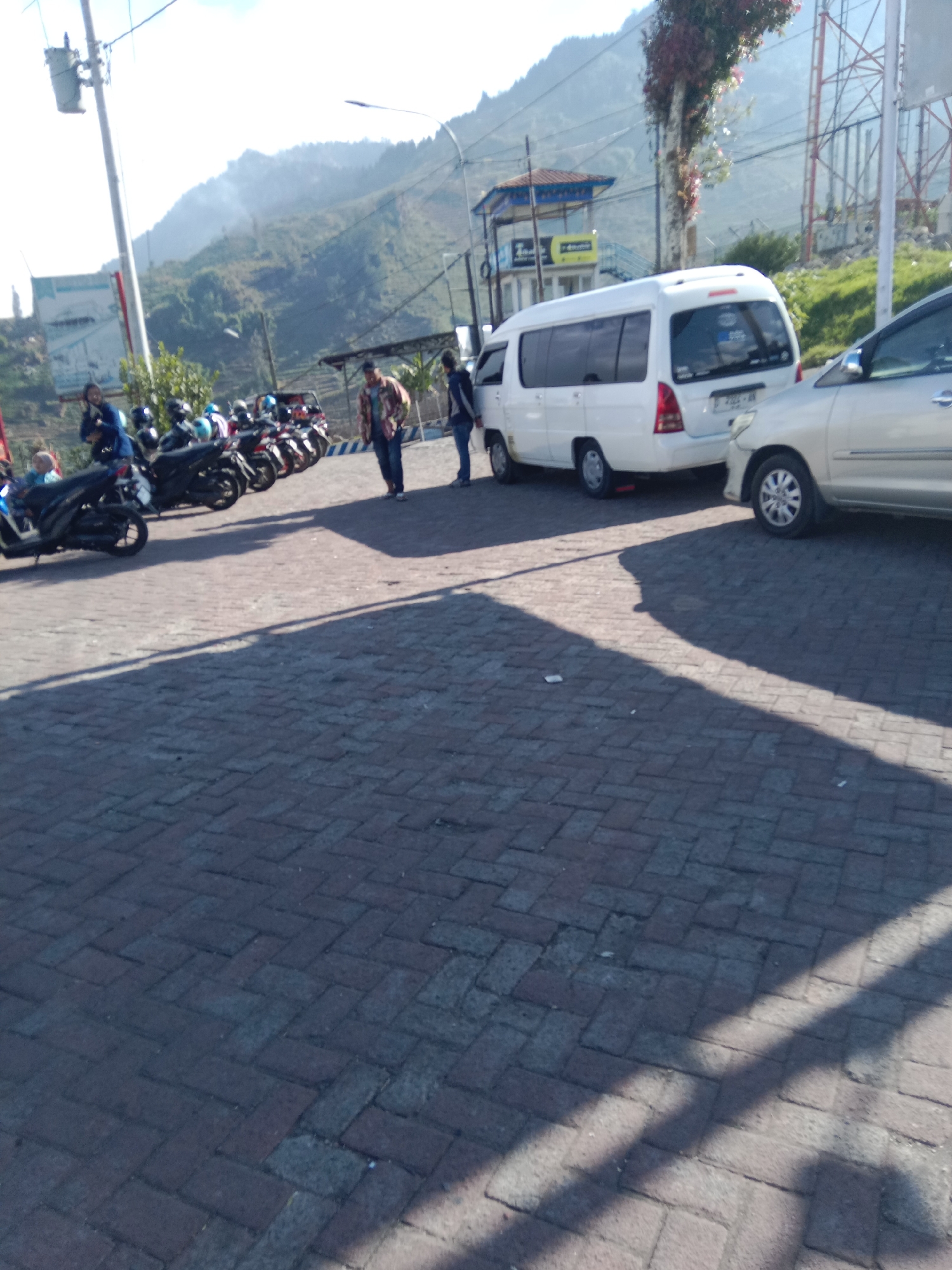 The image captures an outdoor scene in Tieng, Indonesia, characterized by a paved area with brickwork. In the background, a mountainous landscape rises under a bright, possibly hazy, sky. A white minivan is parked in the mid-ground, alongside a silver sedan. Several motorcycles are also visible, suggesting a parking area or a gathering point. Two individuals are standing near the minivan, and another person is seated on one of the motorcycles. The environment appears to be a public space, possibly a viewpoint or a rest stop given the presence of vehicles and the scenic mountainous backdrop. The lighting indicates it is daytime, with strong shadows cast across the brick pavement.